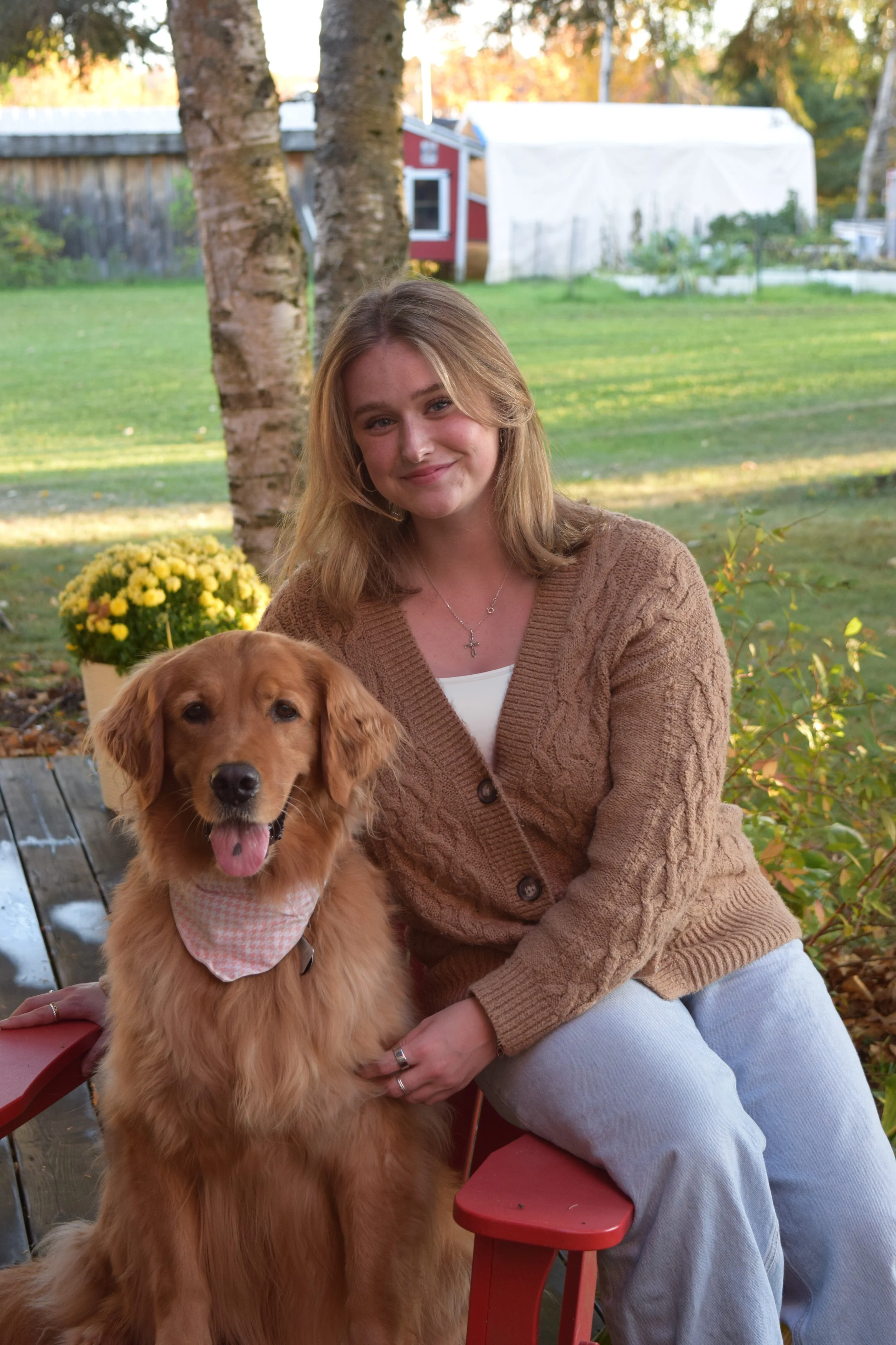A young woman with blonde hair and a brown sweater sitting outdoors with a golden retriever dog on her lap, smiling at the camera. There are trees, a wooden fence, and a garden in the background.