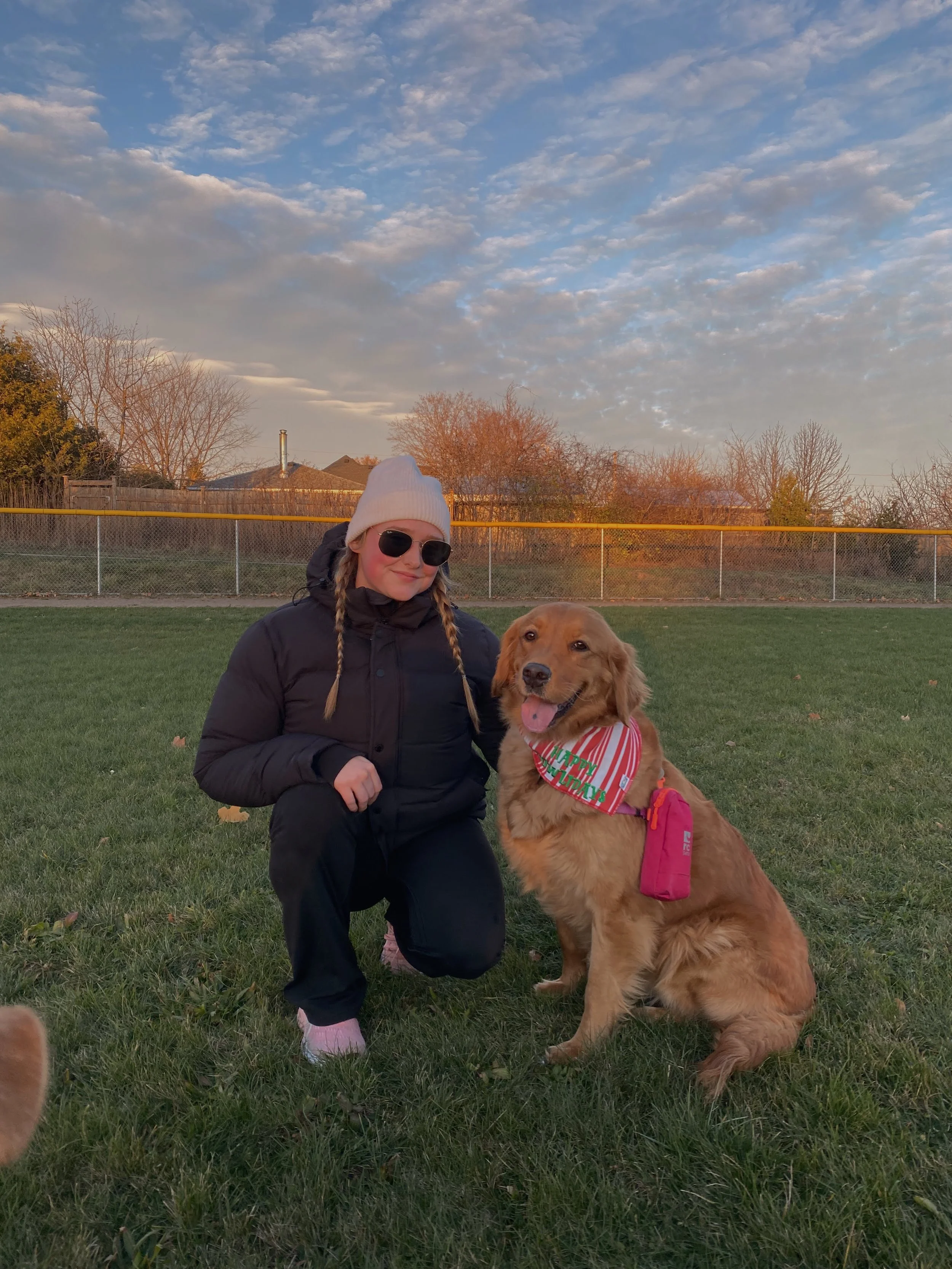 A woman wearing a white knit hat, sunglasses, and dark clothing kneels beside a golden retriever dog with a red and white striped bandana that says "Happy Holidays." They are outdoors on a grassy field with a chain-link fence, trees, and houses in the background under a partly cloudy sky.