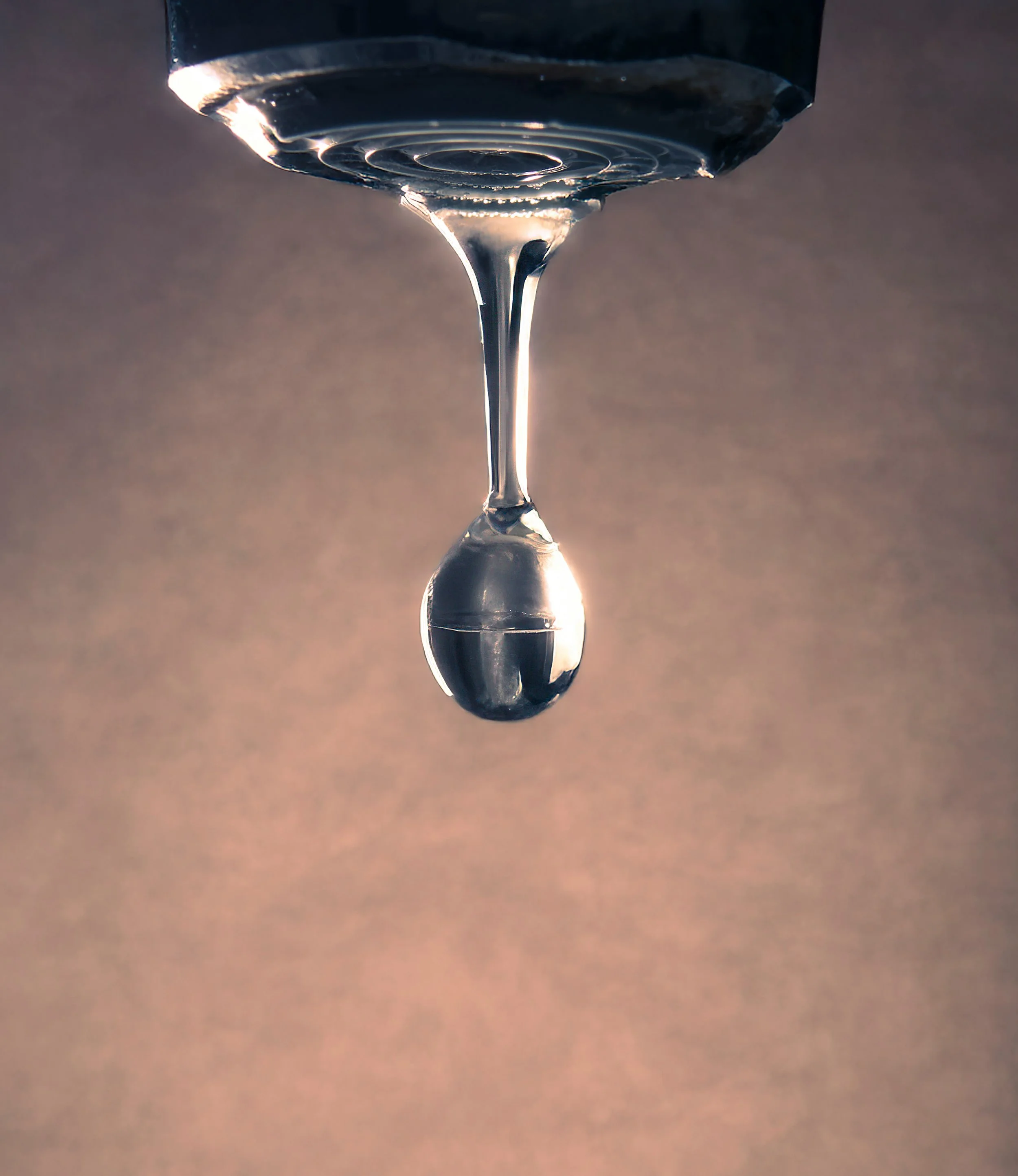 A close-up view of a water droplet hanging from the tip of a faucet, with a blurred background.