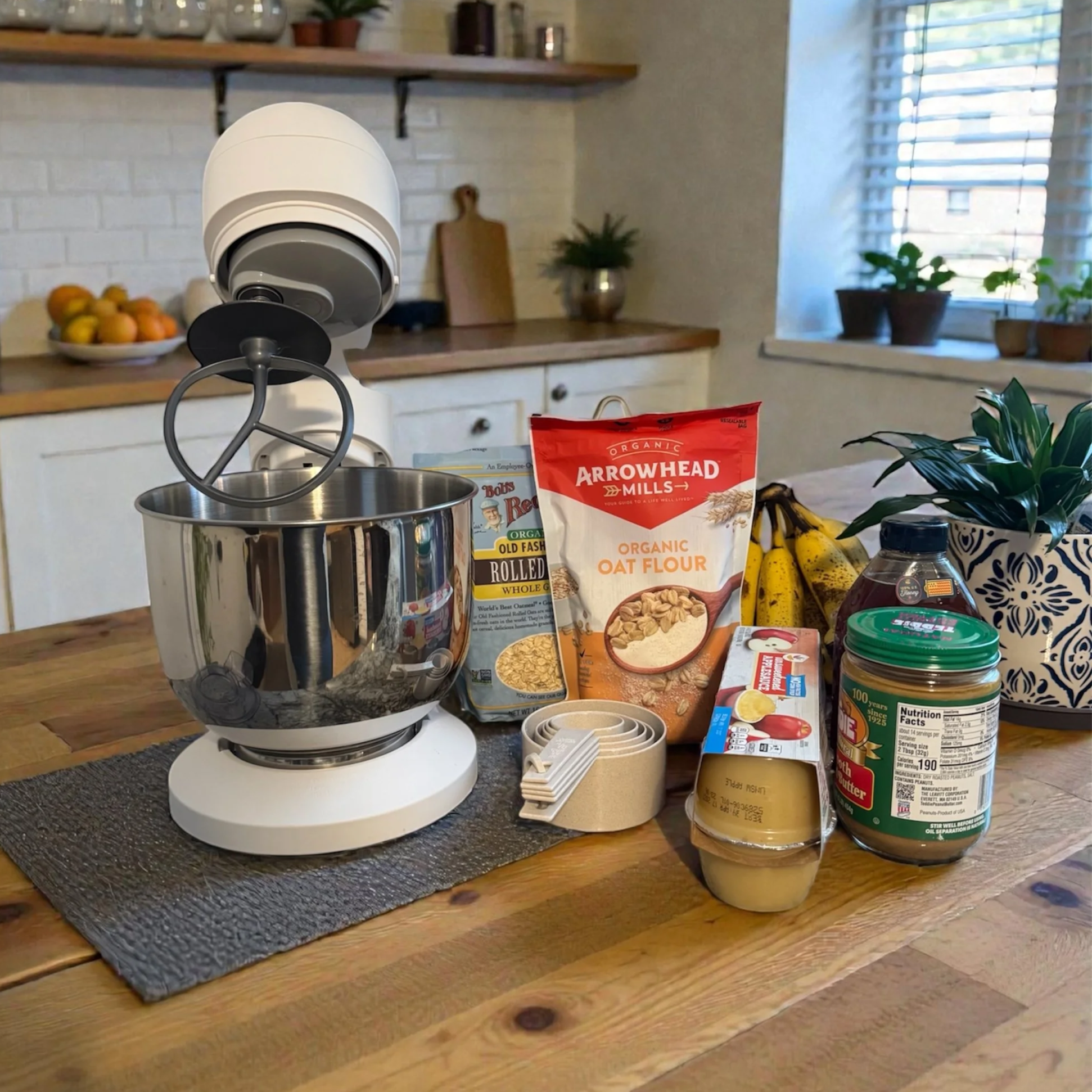 Kitchen countertop with baking ingredients including oat flour, rolled oats, bananas, peanut butter, and measuring cups, with a stand mixer and a plant in a decorative pot.