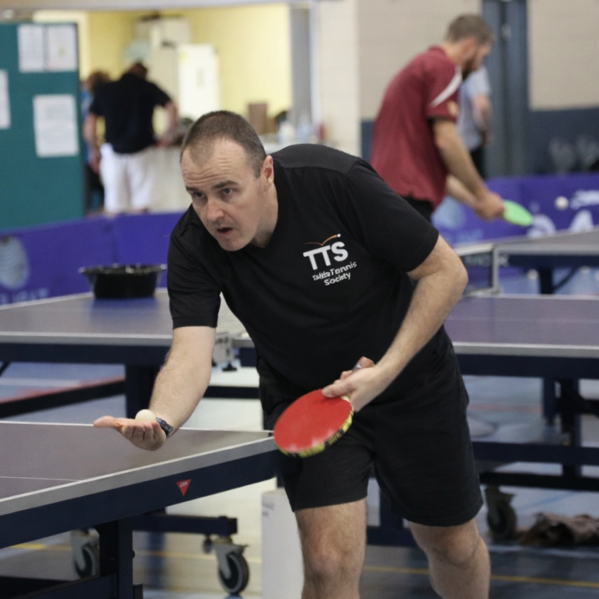A man in a black shirt and black shorts playing ping pong in an indoor gym with other players in the background.