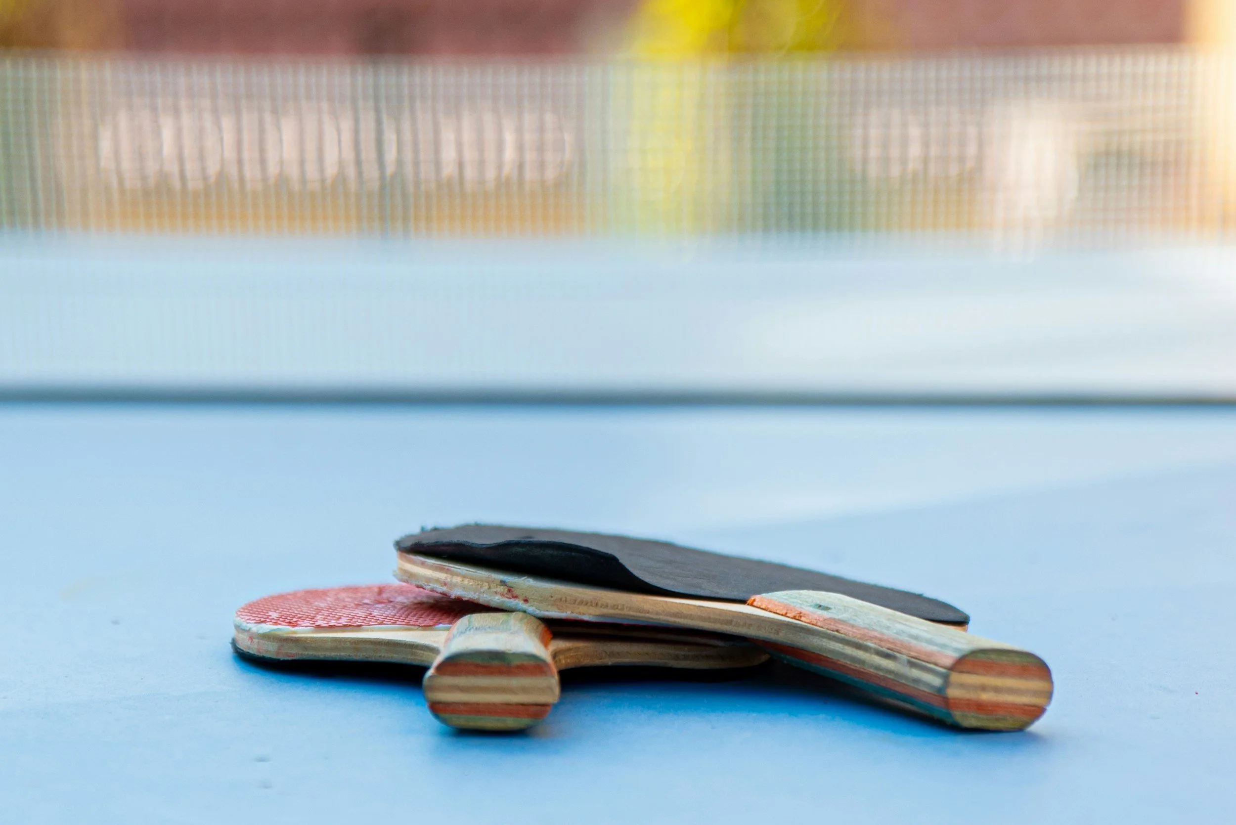 A broken table tennis paddle and ball on a blue table, with a blurry background of a net and outdoor environment.