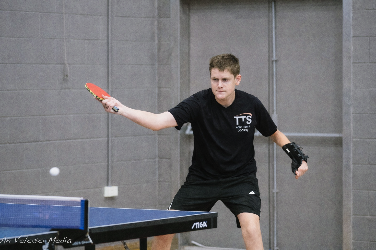 A young man playing table tennis in an indoor gym, wearing a black T-shirt, black shorts, and a black wrist support, focused on hitting the ball.