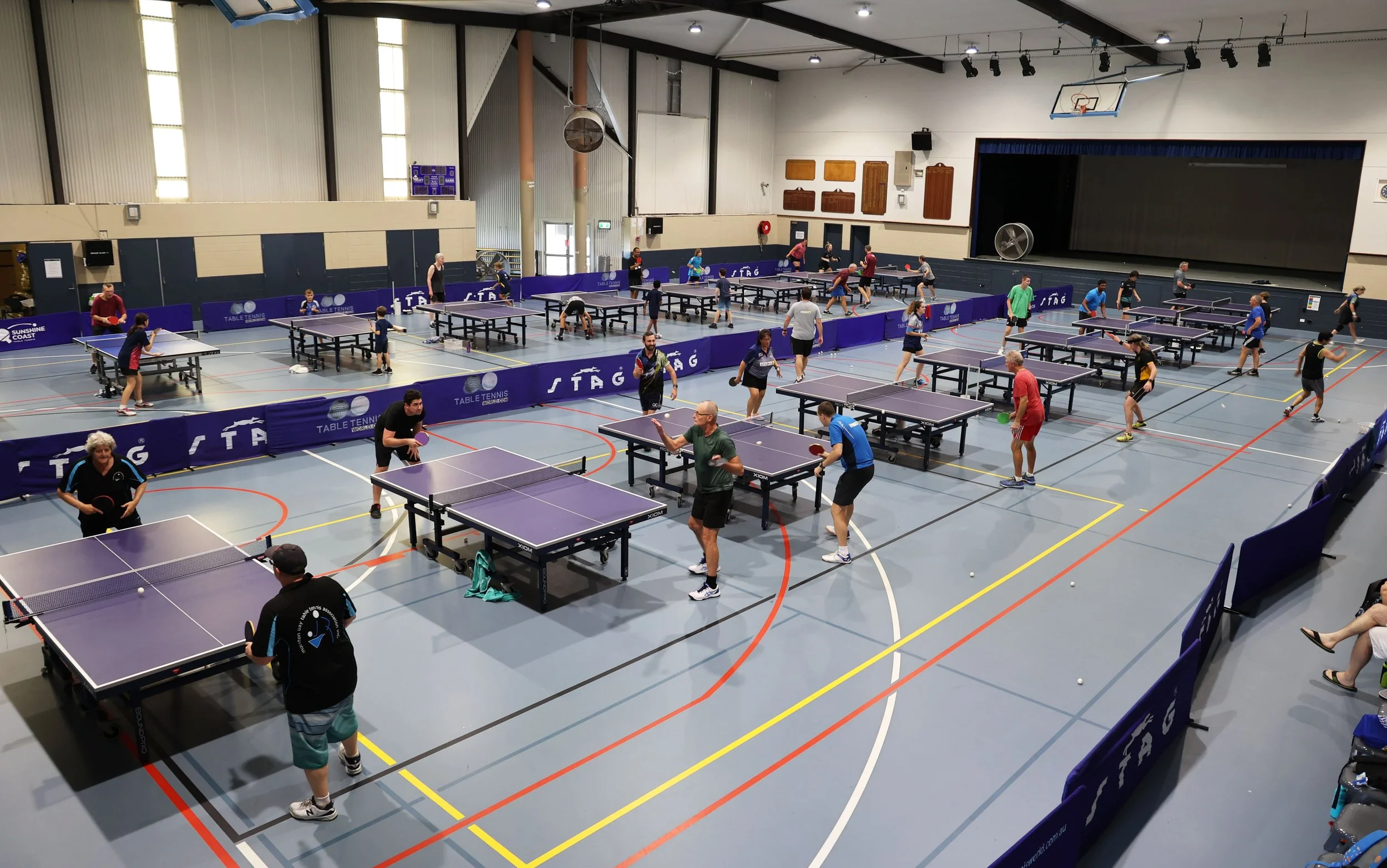 Indoor sports hall with multiple ping pong tables set up for a match or practice, with players of various ages engaged in games, and a stage with a dark curtain at the back of the room.