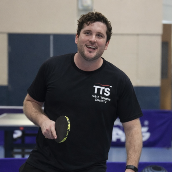 Male table tennis coach smiling in front of table tennis table with bat in right hand,.