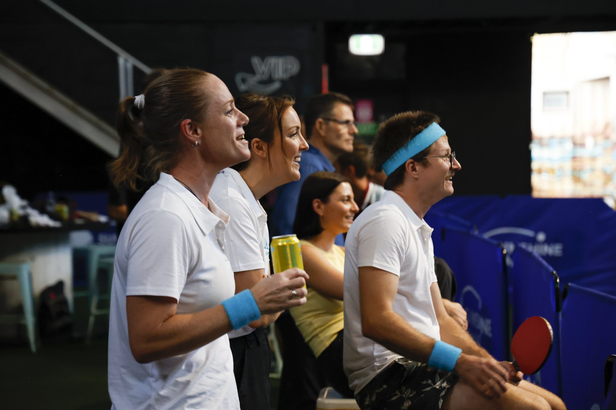 People watching a tennis match, dressed in tennis attire, some holding drinks, sitting and standing in an indoor arena.