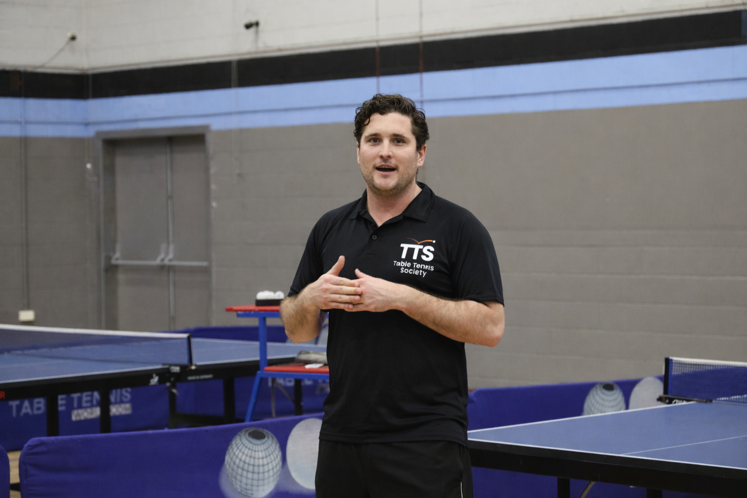 Table Tennis Coach wearing black short with Table Tennis Society logo on it. Speaking to camera with table tennis tables in background.