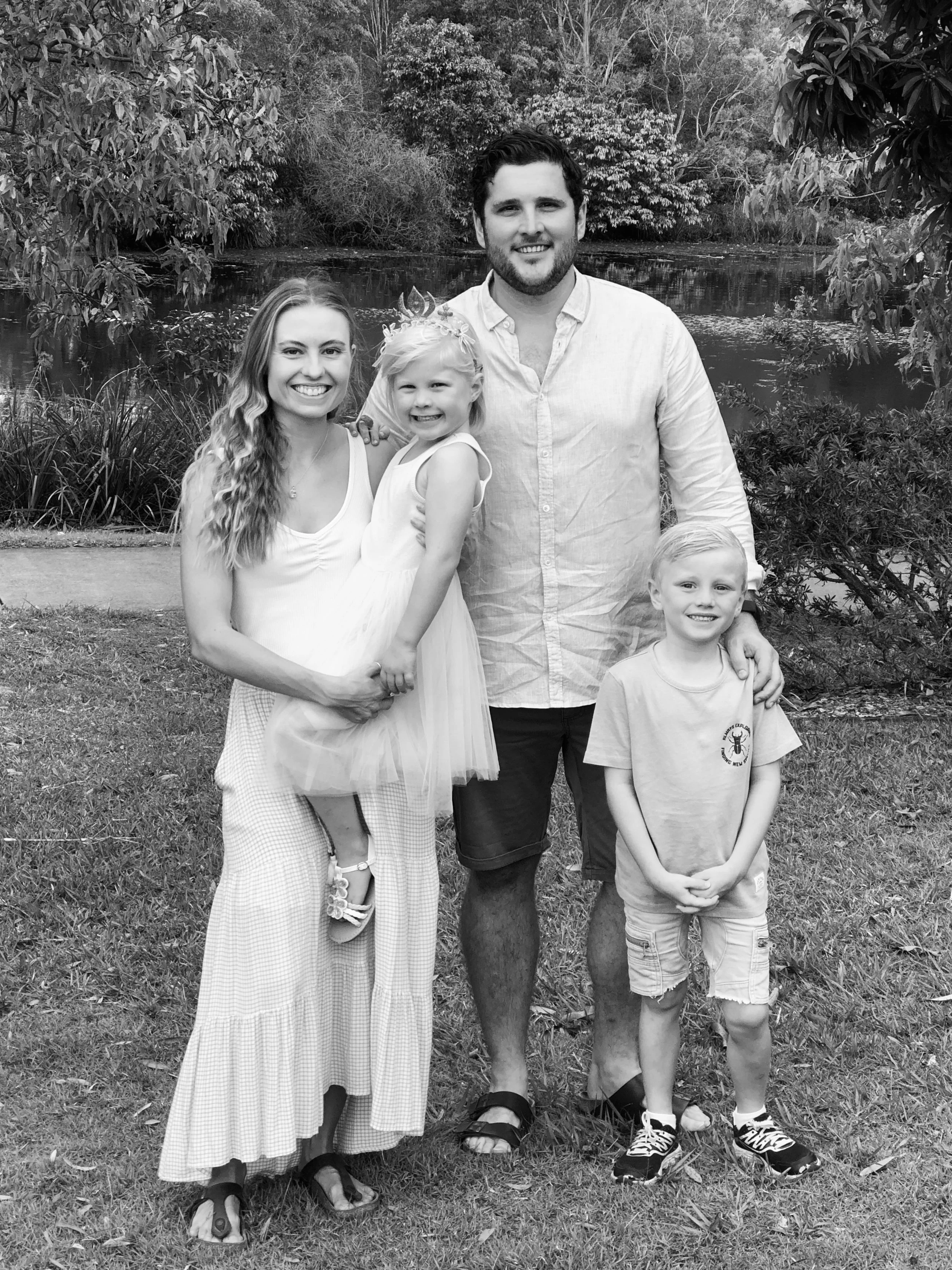 A family of four standing on grass near a lake surrounded by trees, smiling at the camera. The mother holds a young girl in a dress, and the father stands next to a boy in shorts.