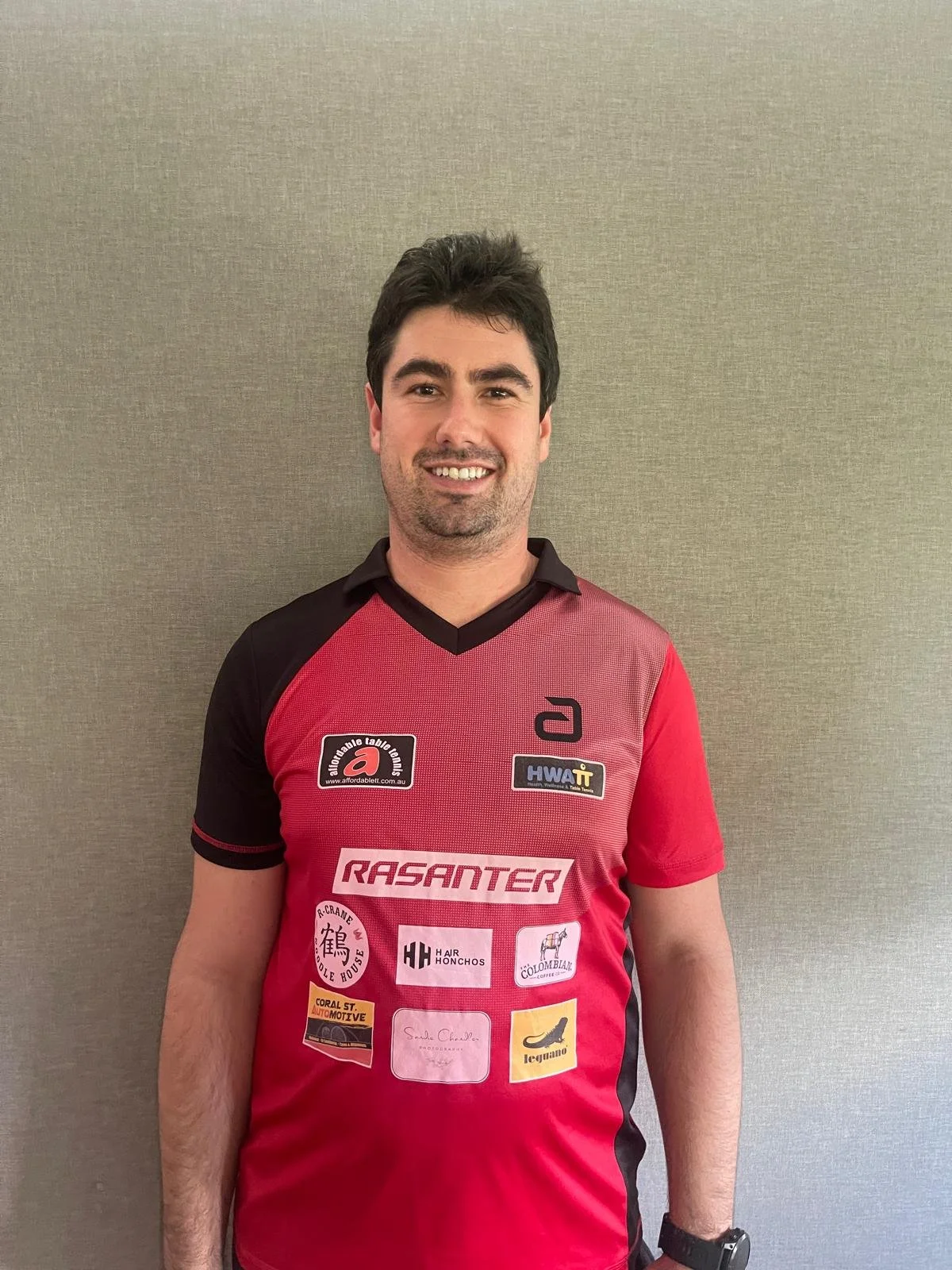 A man with dark hair and a beard wearing a red and black sports jersey, standing against a beige wall, smiling at the camera.