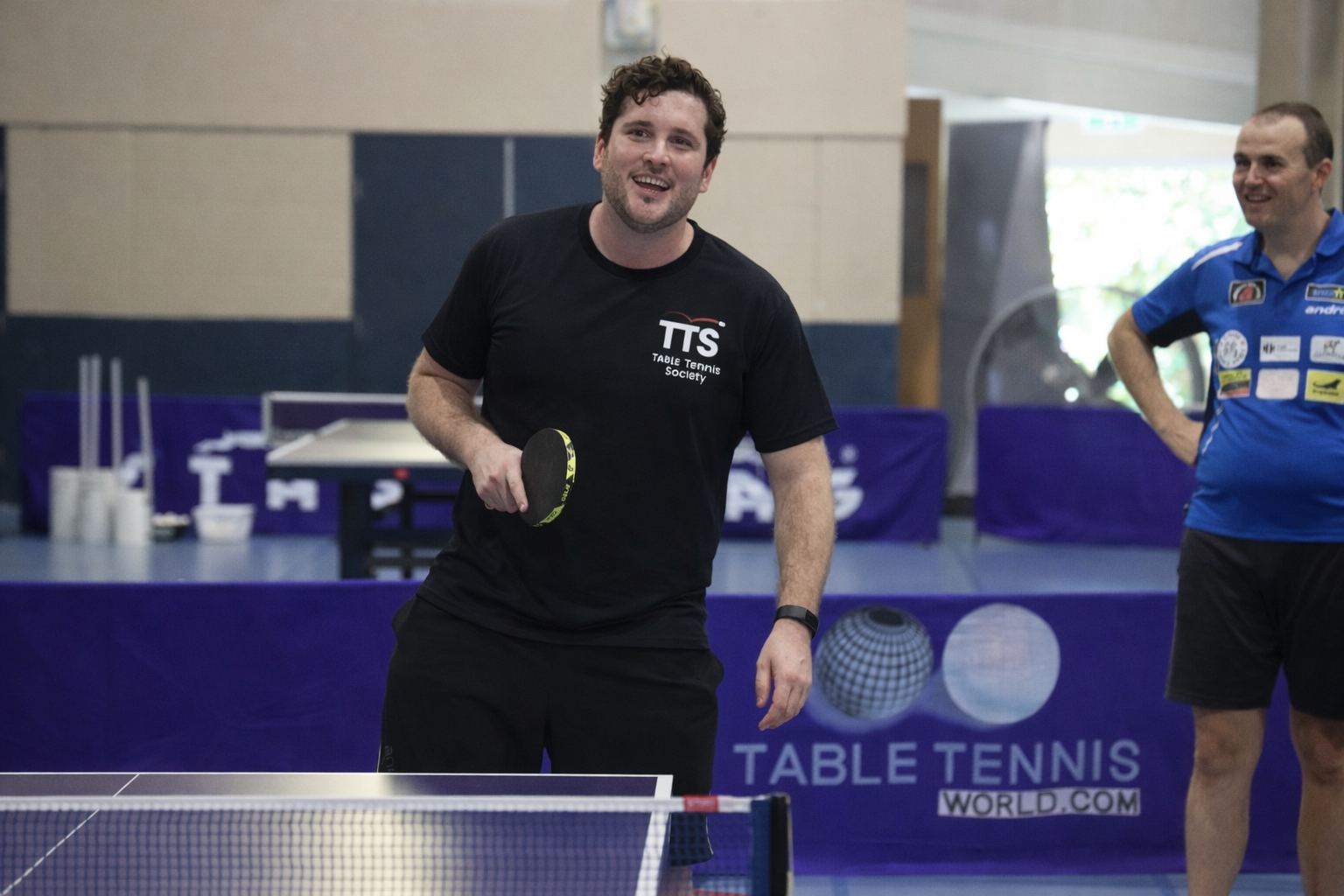 Male Coach smiling with table tennis bat in right hand and black shirt. Standing in front of a table tennis table,