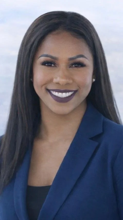 Close-up of a smiling woman with dark hair, wearing a blue blazer and dark lipstick, standing outdoors with a blurred ocean background.
