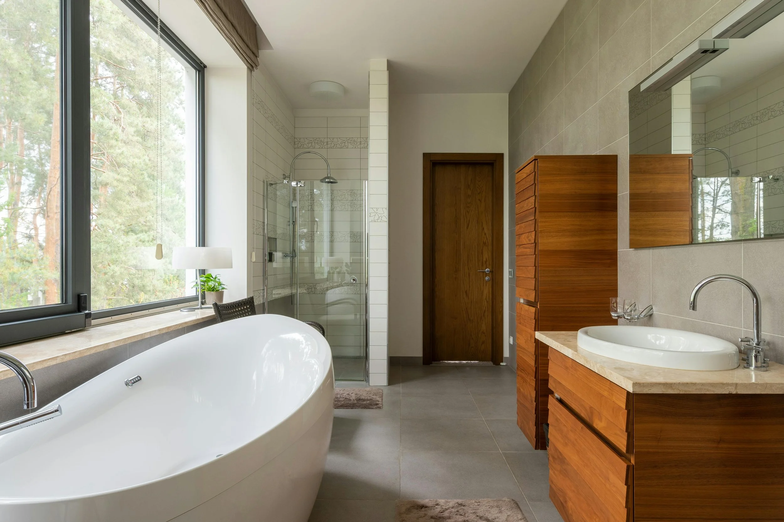 Modern bathroom featuring a large oval bathtub near a window, a walk-in shower with glass doors, a wooden cabinet, and a wooden vanity with a round sink, with a large mirror and neutral color tones.