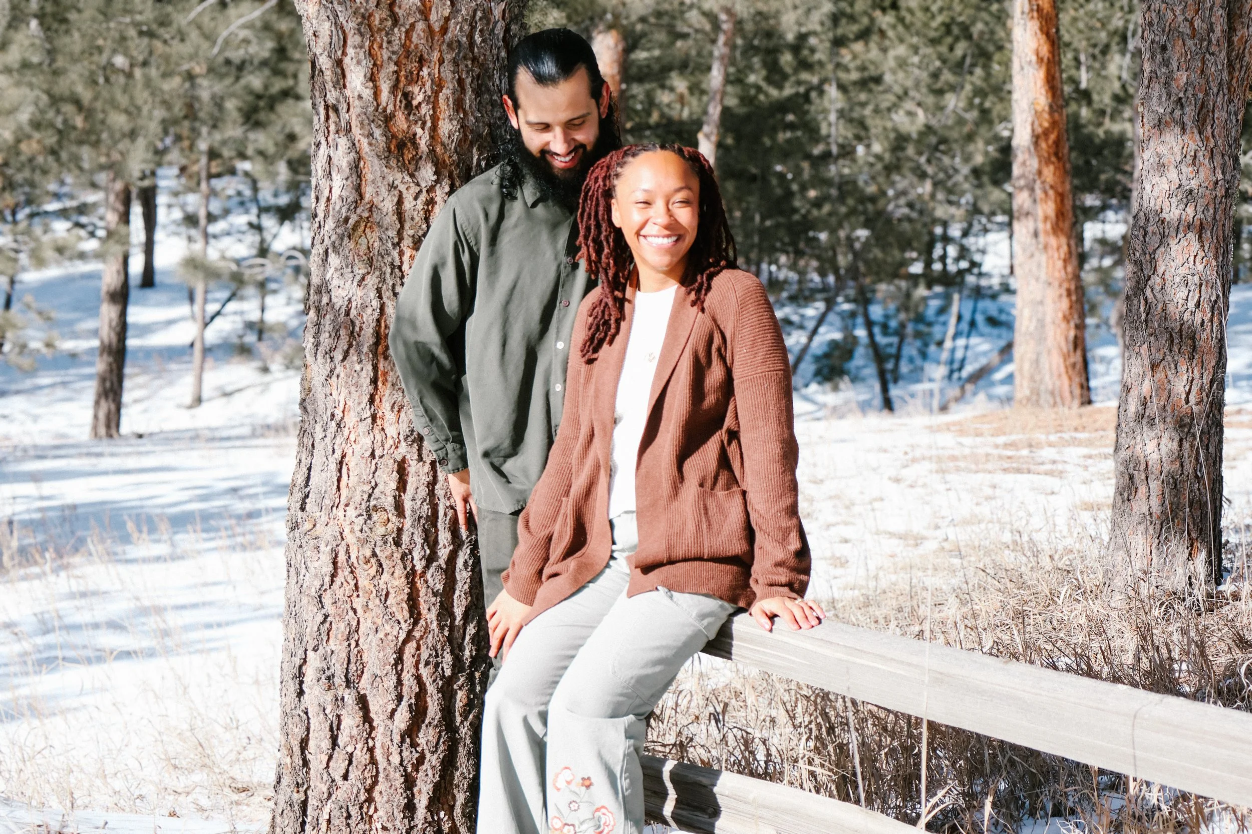 A man and a woman enjoy a snowy outdoor setting with trees, sitting on a wooden fence, smiling and laughing.
