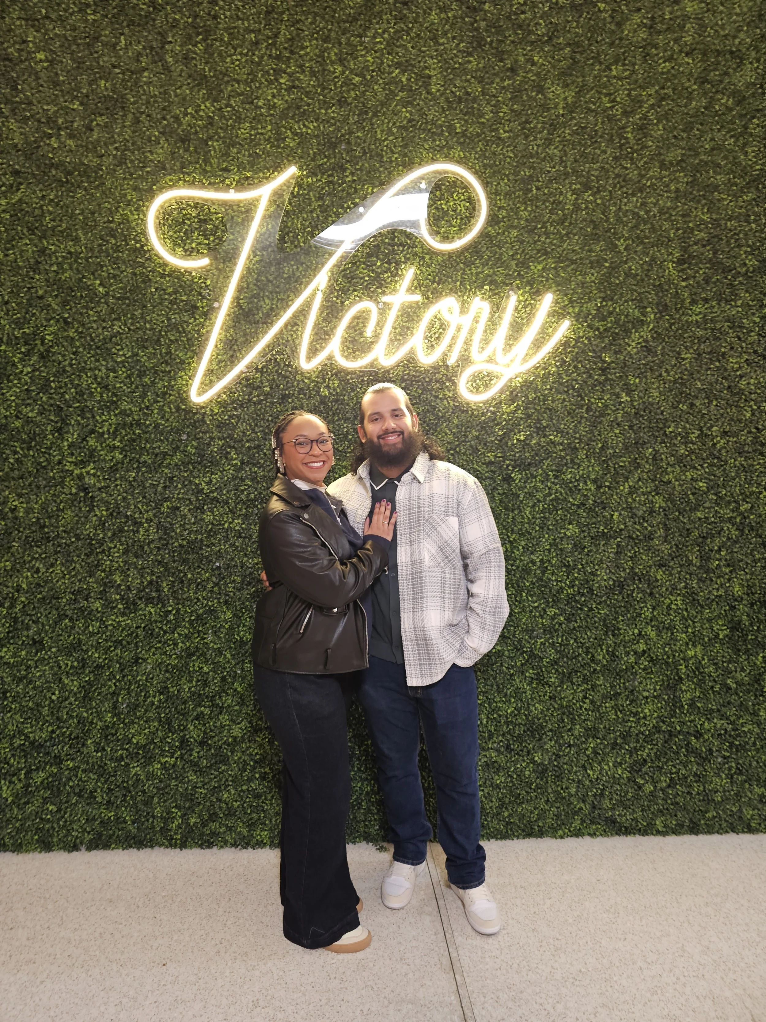 A smiling couple standing in front of a green wall with a neon sign that says 'Victory' and a cocktail glass design. The woman is wearing a black leather jacket and glasses, and the man is wearing a gray plaid jacket.