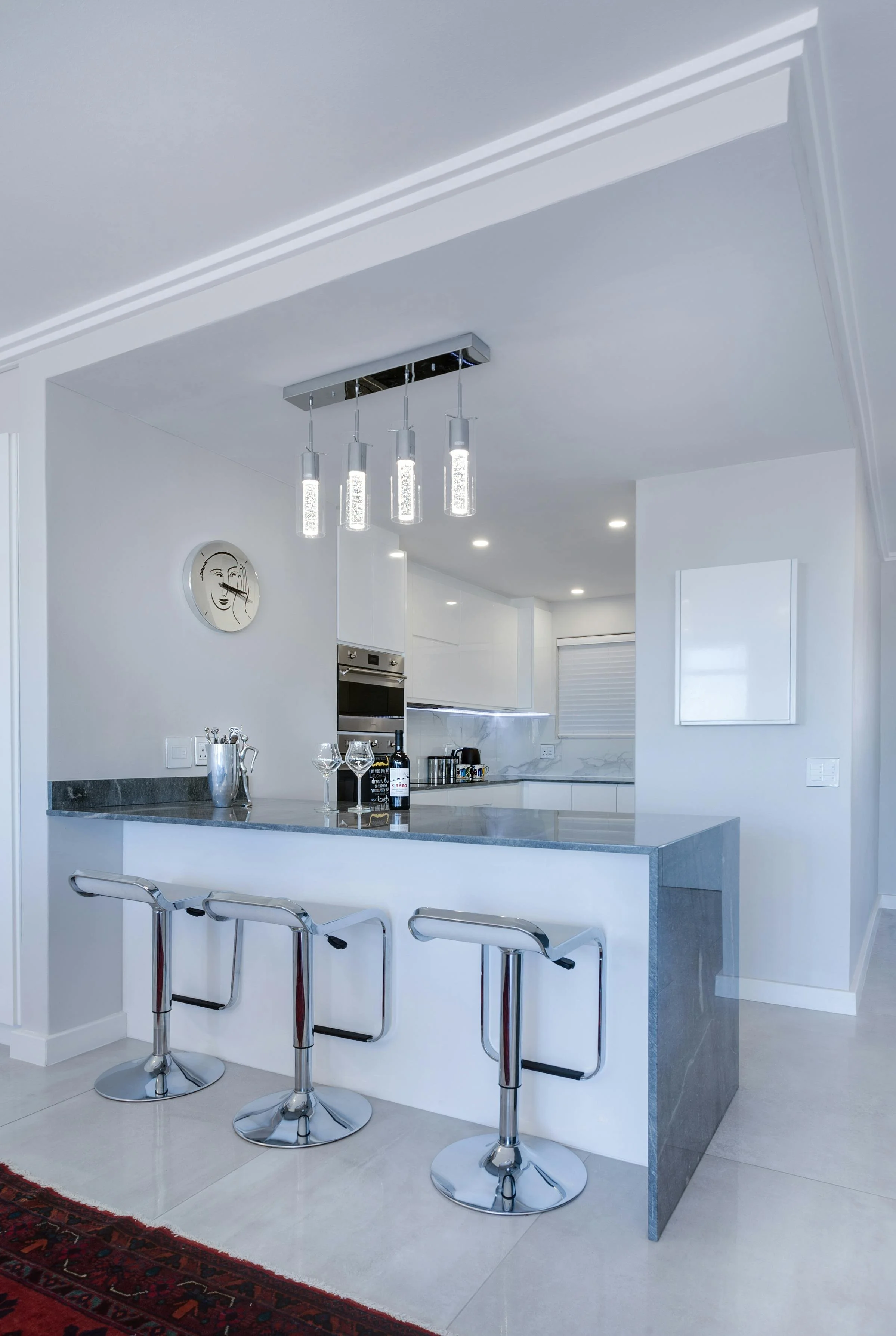 Modern white kitchen with a gray countertop island, three chrome bar stools, and decorative pendant lighting, with a wall clock.
