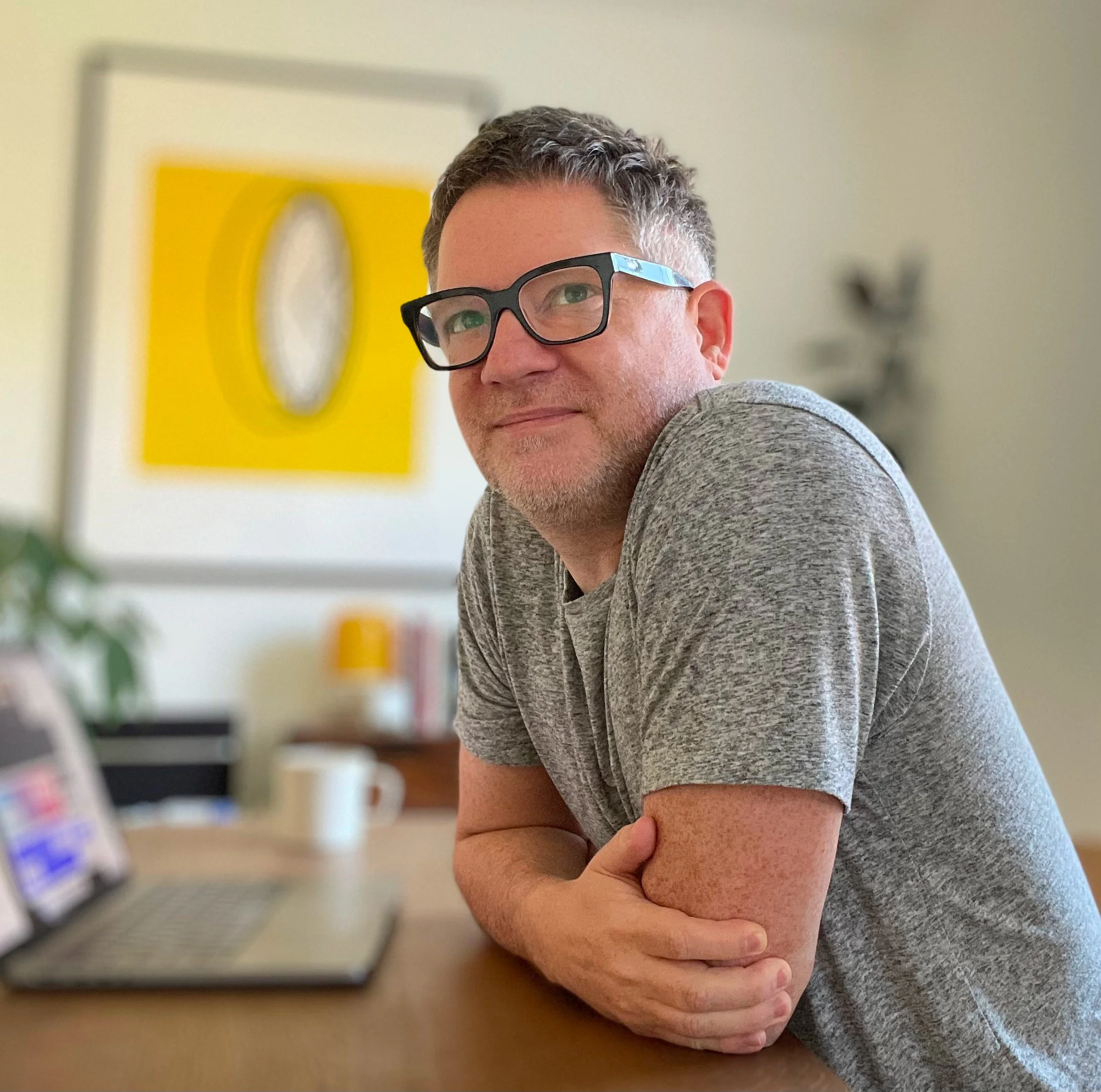 A man with glasses, gray hair, and a beard, resting his head and arm on a person's lap in a room with a yellow and gray wall and shelves with products.