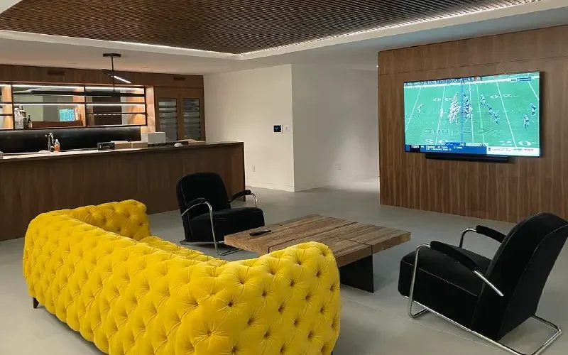 Modern living room with a large flat-screen TV showing a football game, a yellow tufted sofa, three black armchairs, a wooden coffee table, a bar area in the background, and a wooden ceiling panel.