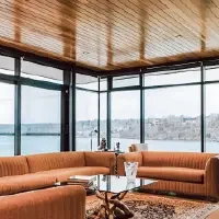 Modern living room with large glass windows overlooking a river and landscape, featuring pink sofas, a glass coffee table, and decorative books and sculptures.