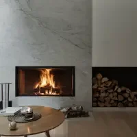 Modern living room with a white marble fireplace, and a wooden ceiling. There is a round wooden table with a candle, a remote, and a ball on it, and a log holder with firewood.