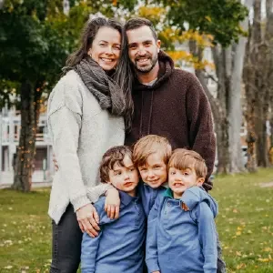 A family of five smiling together outdoors in a park during autumn, with trees and fallen leaves in the background.