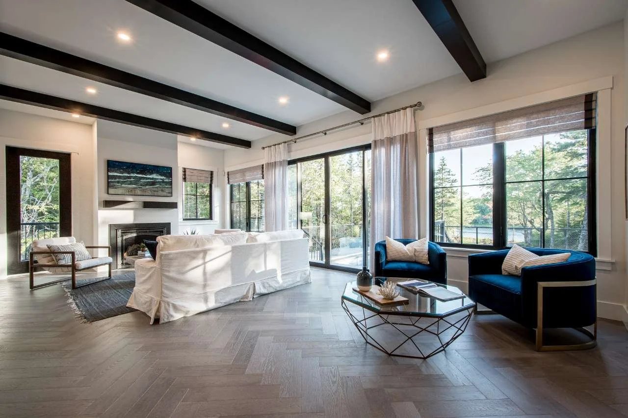 Bright living room with white sofa, two blue armchairs, a glass coffee table, and large windows showing greenery outside.
