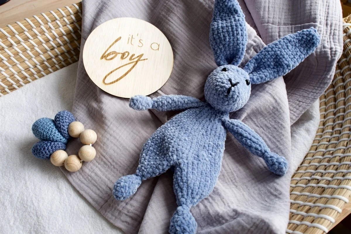 Een zachte, blauwe knuffelkonijn ligt op een zacht doekje in een houten wieg. Onder het konijn ligt een ronde houten muurdecoratie met de tekst 'it's a boy'. Daarnaast ligt een kralenring van hout en blauwe knoopjes, mogelijk voor een baby. De setting is warm en knuffelig, typisch voor een babycouture.
