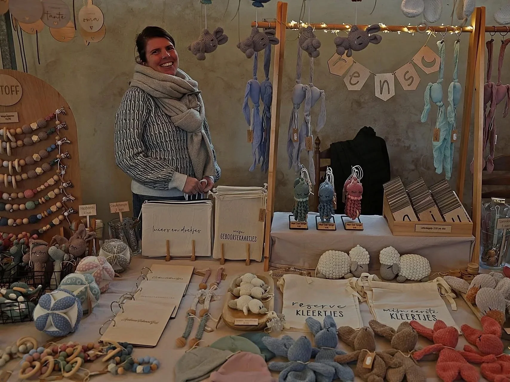 Vrouw die op een marktkraam met handgemaakte knuffels en accessoires staat, met houten hangers met gebreide knuffels en kaarten, en een verscheidenheid aan gehaakte en houten sieraden.
