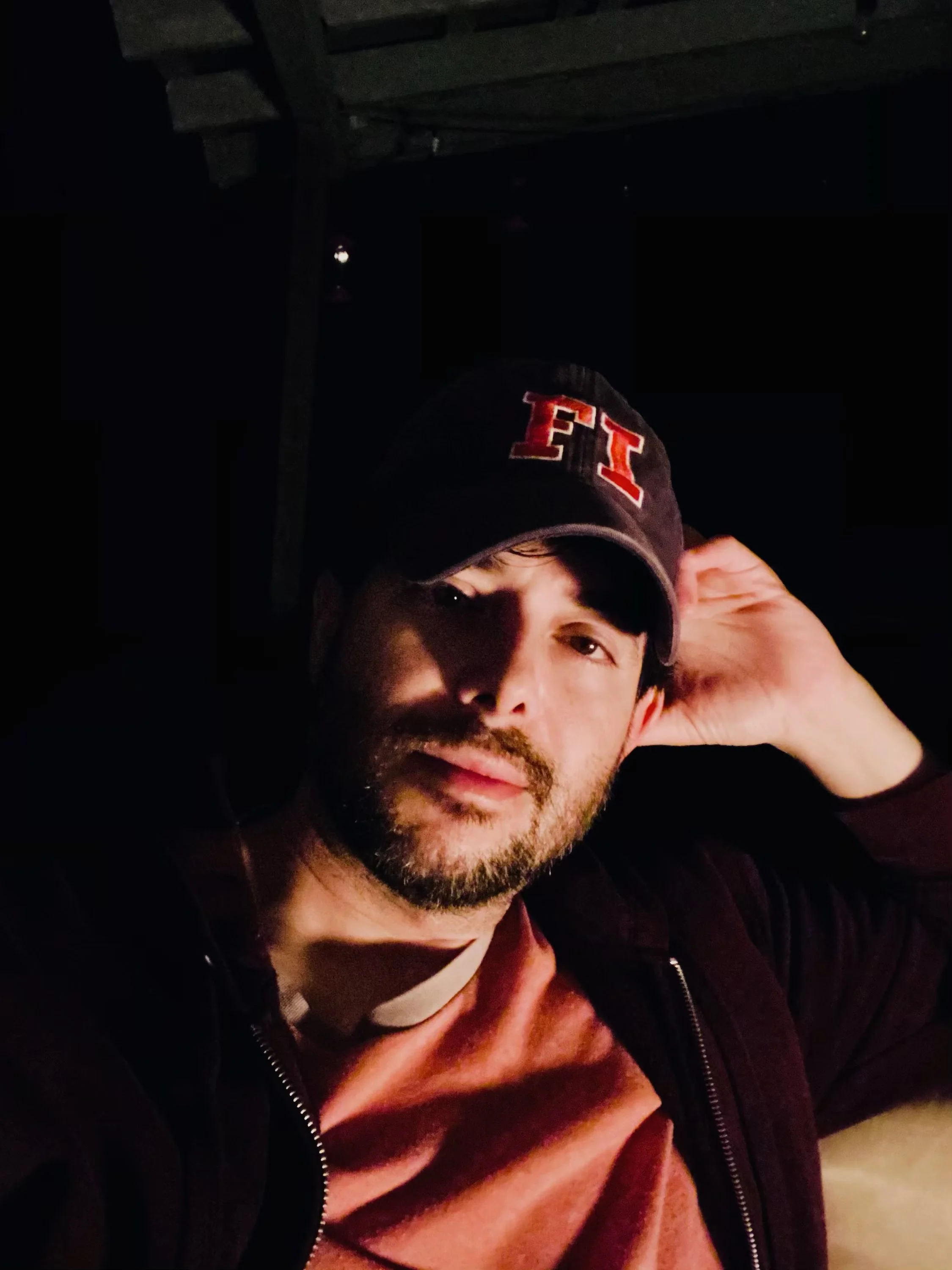 A man with a beard wearing a black cap with red and white lettering, a black jacket, and a reddish shirt, sitting in a dimly lit environment, looking at the camera with one hand behind his head.