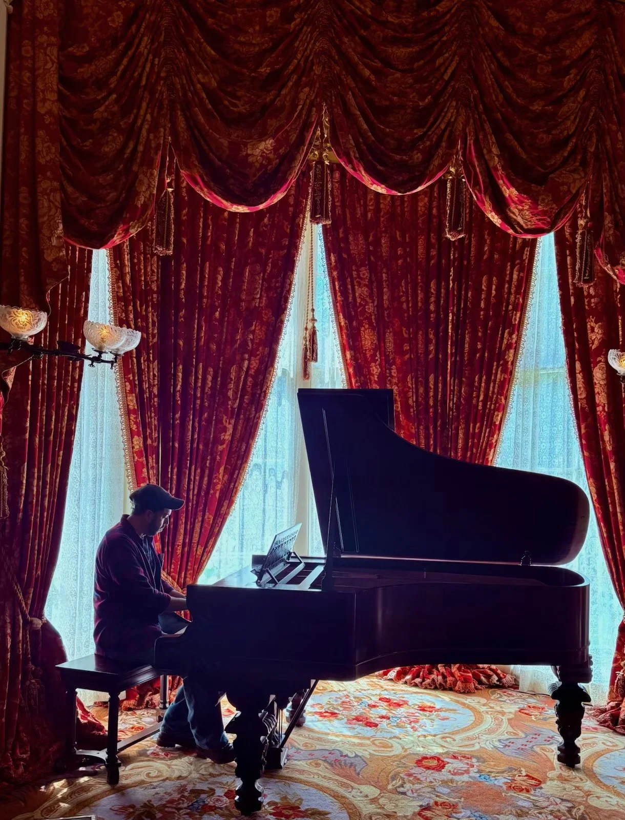 A person sitting at a grand piano in a room with red patterned curtains and sheer blue curtains, with ornate floral carpet on the floor.
