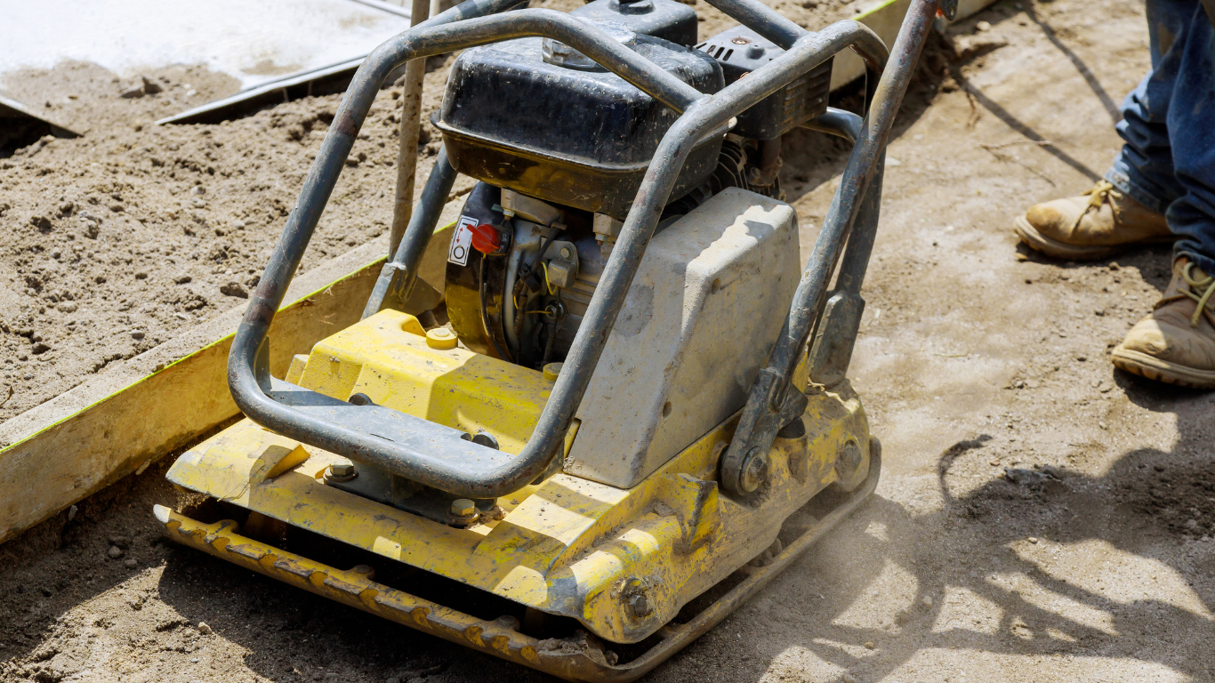 A yellow plate compactor on a construction site with dirt and sand, surrounded by the legs of a person wearing work boots and blue jeans.
