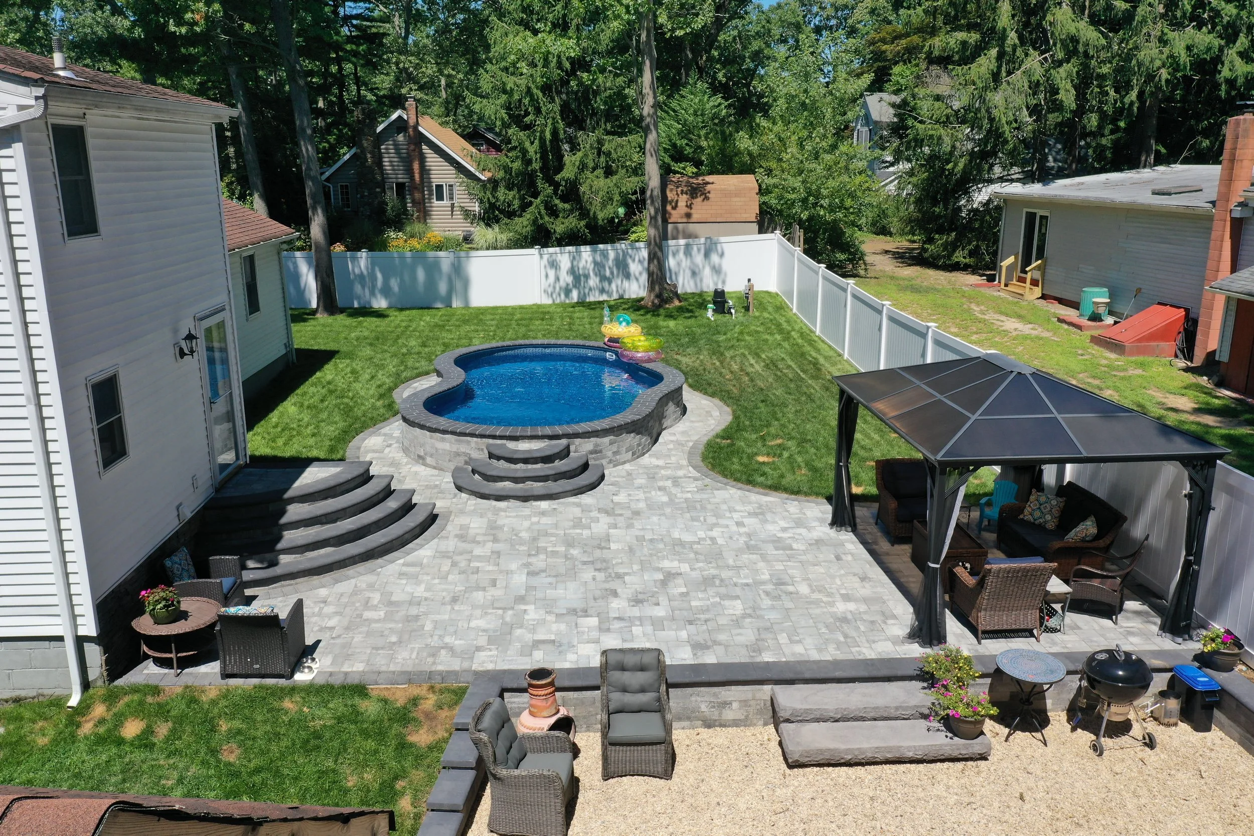 A backyard with a kidney-shaped swimming pool on a patio made of grey pavers, surrounded by a white fence and green grass. There is a set of curved steps leading to the pool, and various outdoor furniture including a gazebo with seating, a small round table, chairs, and a barbecue grill.