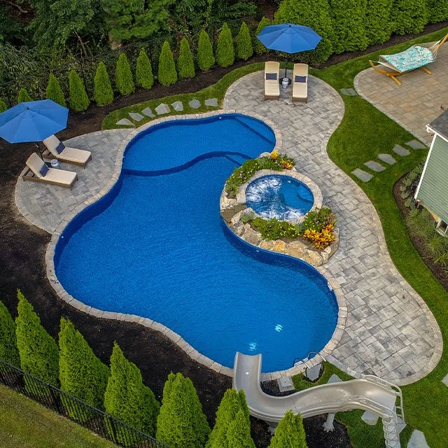 A backyard with a kidney-shaped swimming pool, a hot tub, a waterslide, patio furniture, umbrellas, and manicured landscaping.