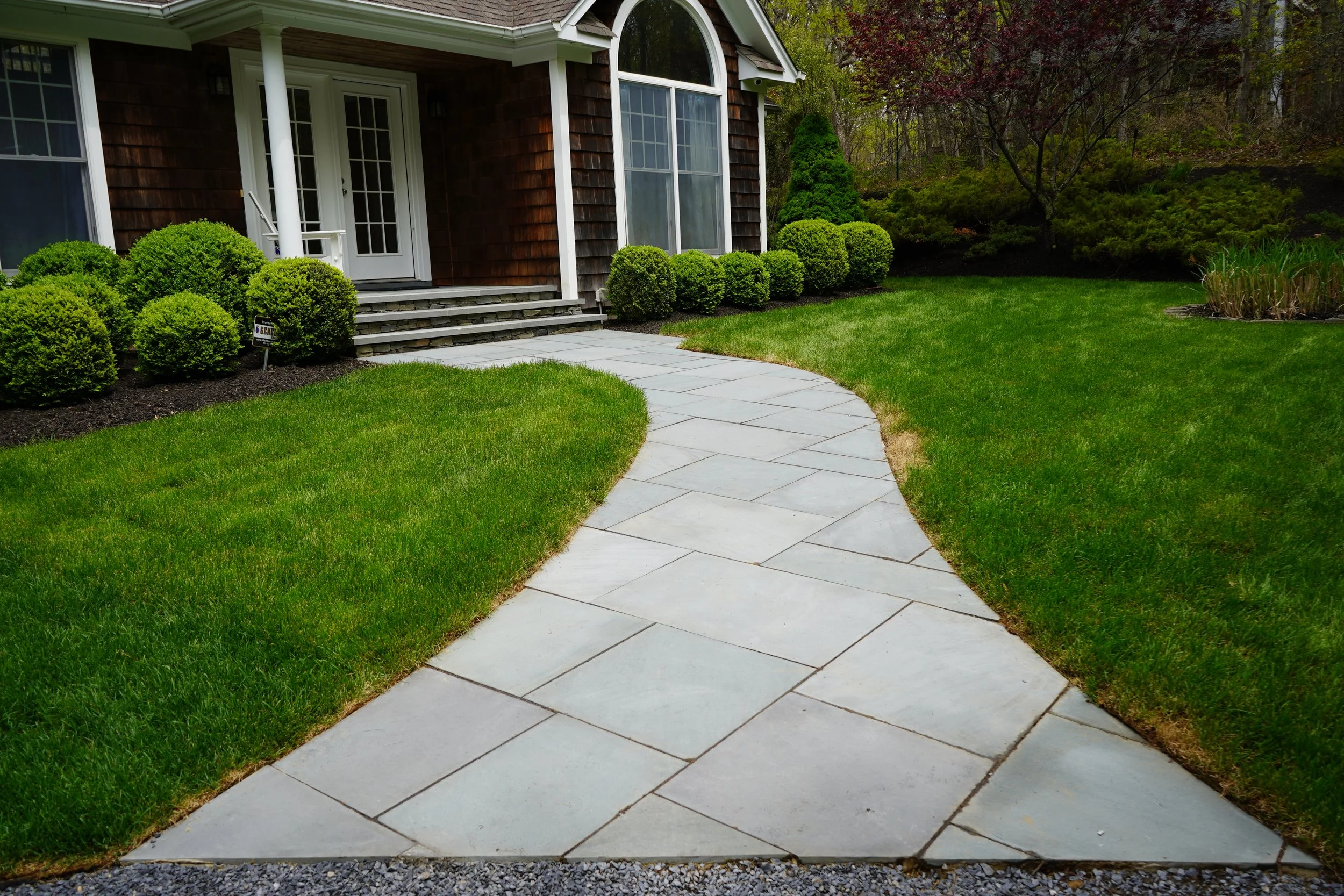 A curved concrete walkway in front of a house with wooden siding, white doors, and large windows, surrounded by a well-maintained lawn and neatly trimmed shrubs.