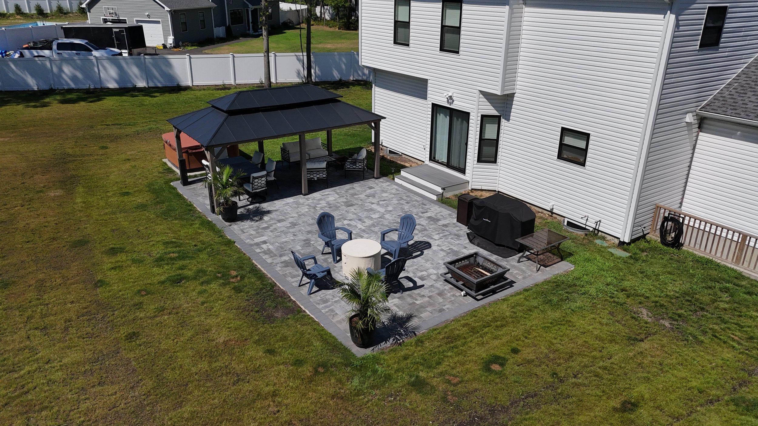A backyard patio with a covered seating area, outdoor dining table with chairs on a gray brick surface, a barbecue grill, fire pit, and a few potted plants, surrounded by a grassy lawn and a white house in the background.