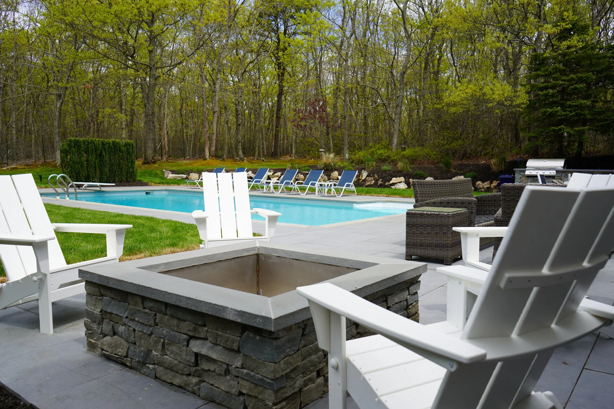 Residential backyard with a swimming pool surrounded by lounge chairs, outdoor seating, and a barbecue grill, with a wooded area in the background.