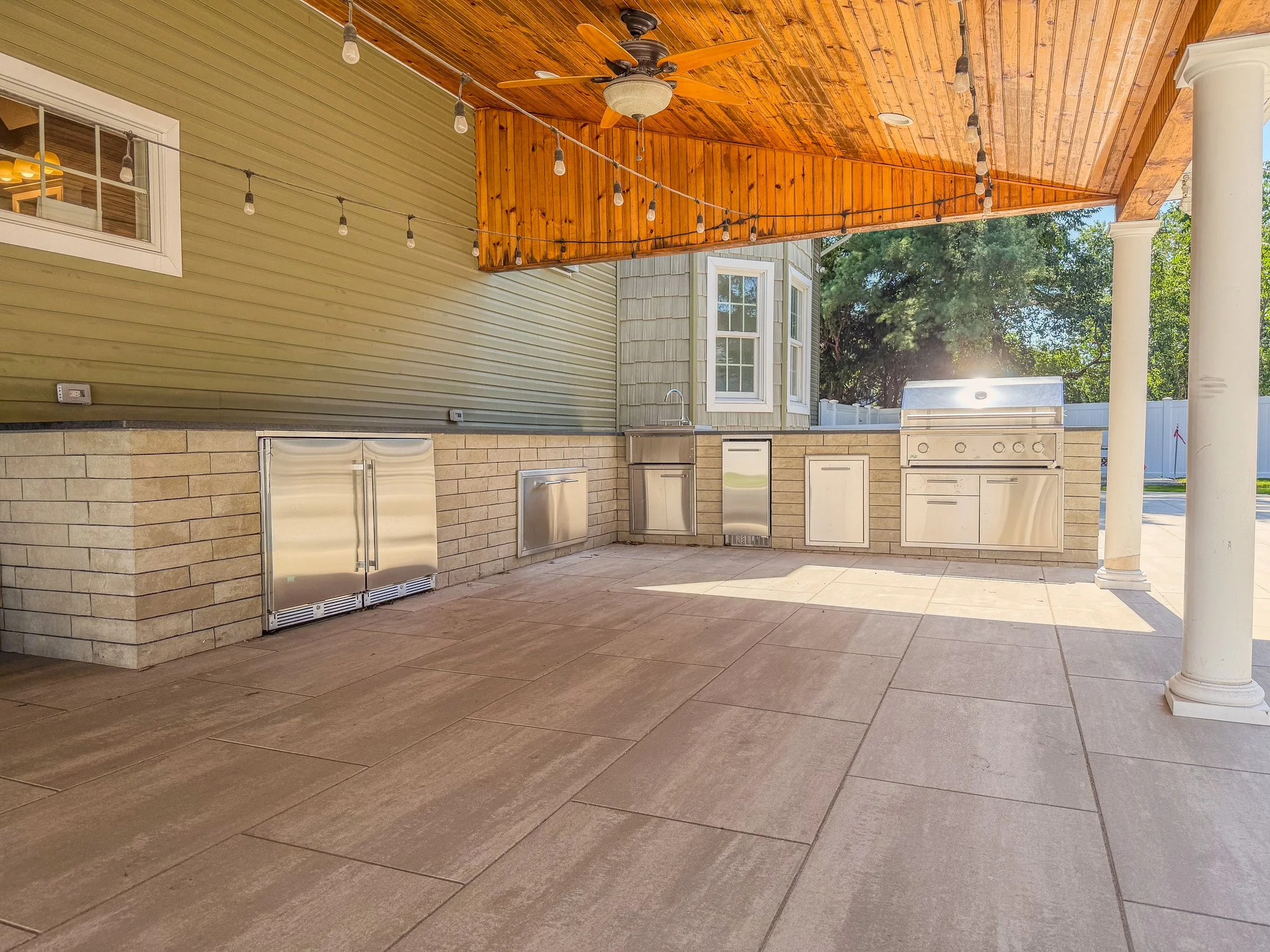 Outdoor kitchen with stainless steel appliances under a wooden covered patio with string lights and a ceiling fan, surrounded by a beige brick wall and columns.