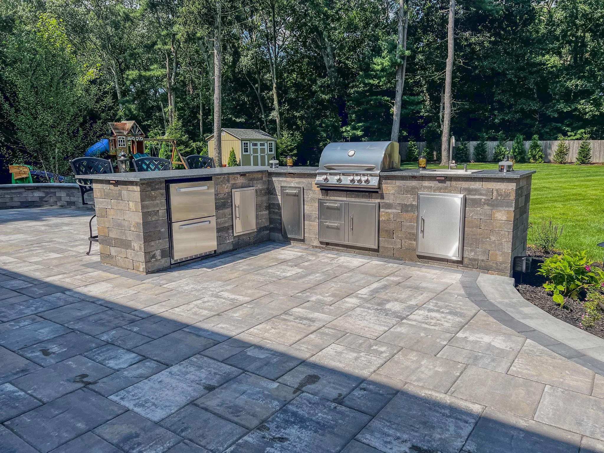 Outdoor kitchen with brick and stainless steel appliances, including a grill, refrigerator, and cabinets, situated on a paved patio in a backyard with grass, trees, a shed, and a playground in the background.