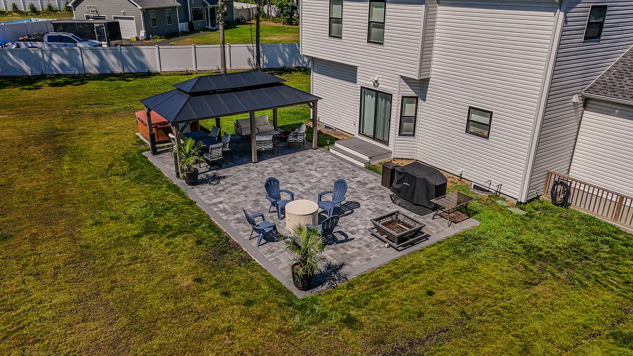 A backyard patio with outdoor furniture, a fire pit, a grill, and a gazebo next to a white house with a sliding glass door, surrounded by a grassy area and fencing.