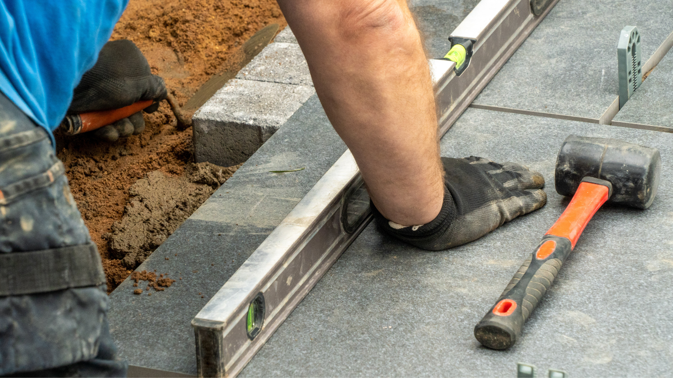 A construction worker installing patio tiles, using a level, with a rubber mallet nearby.