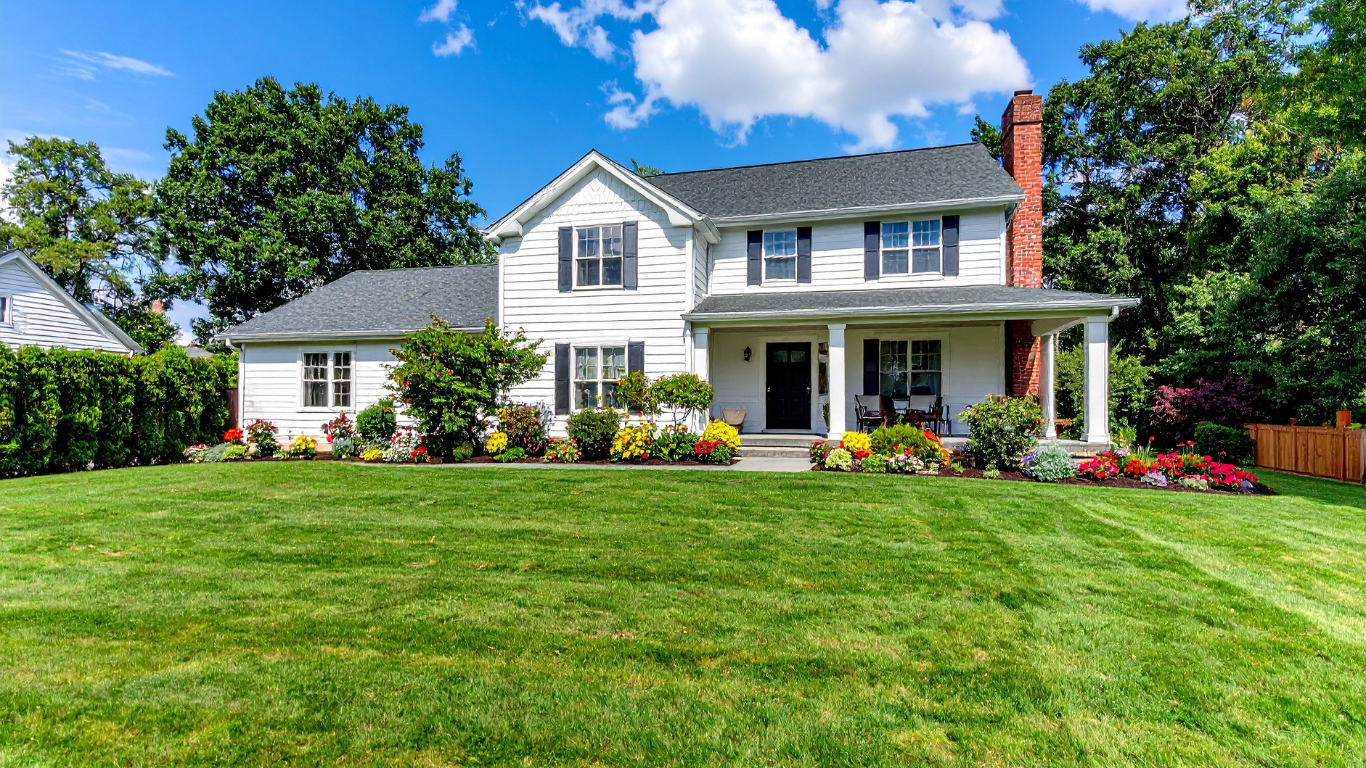 White two-story house with black shutters, a covered front porch, and a manicured lawn with colorful flower beds, trees, and a partly cloudy blue sky.