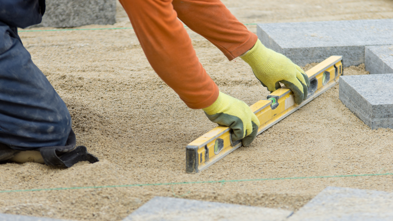 Person laying stones with a spirit level on a sandy surface at a construction site.