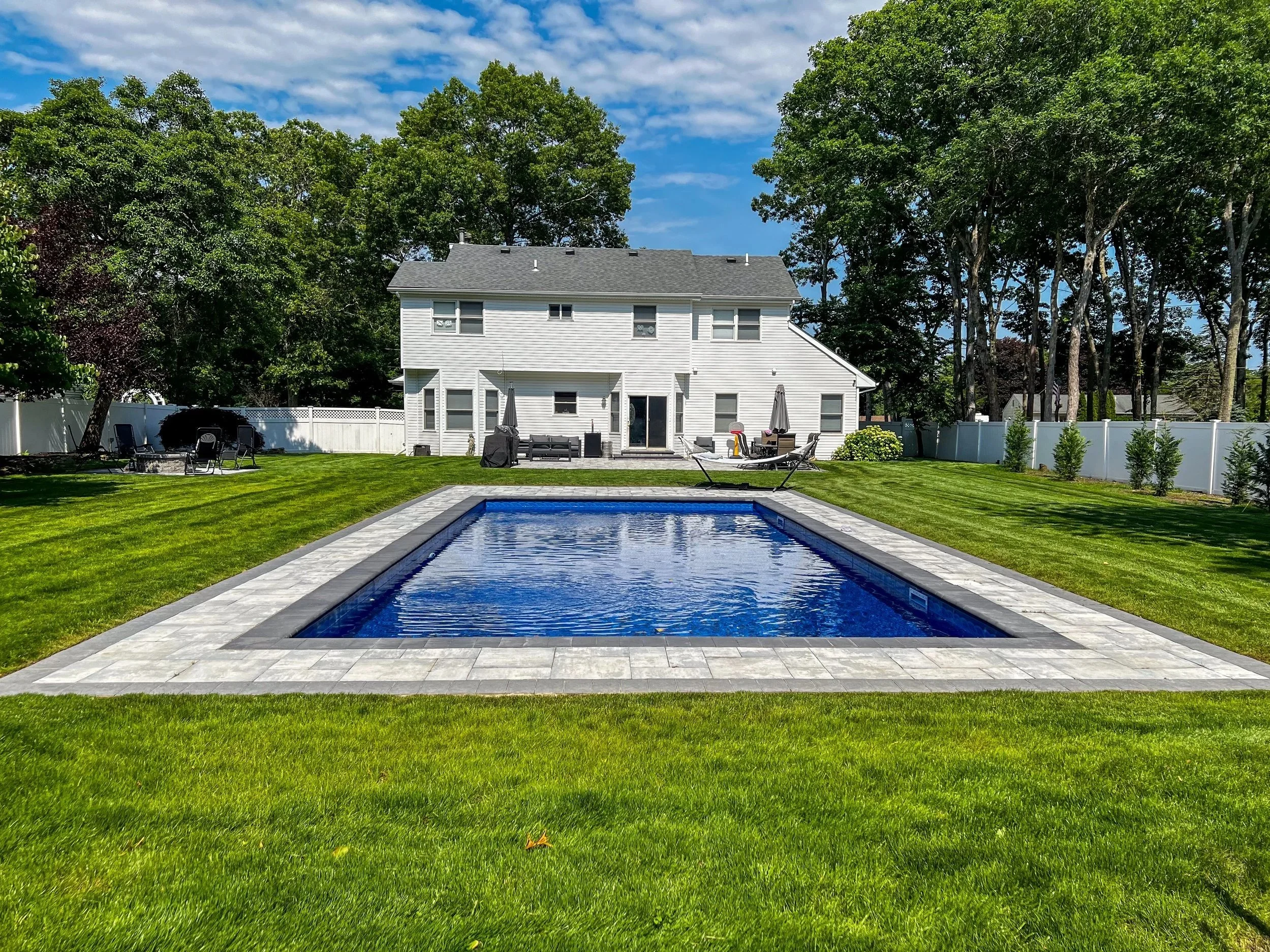 Backyard with a rectangular swimming pool, paver patio around the pool, green grass, white house with outdoor furniture, BBQ grill, lounge chairs, umbrellas, surrounded by trees and a white fence.