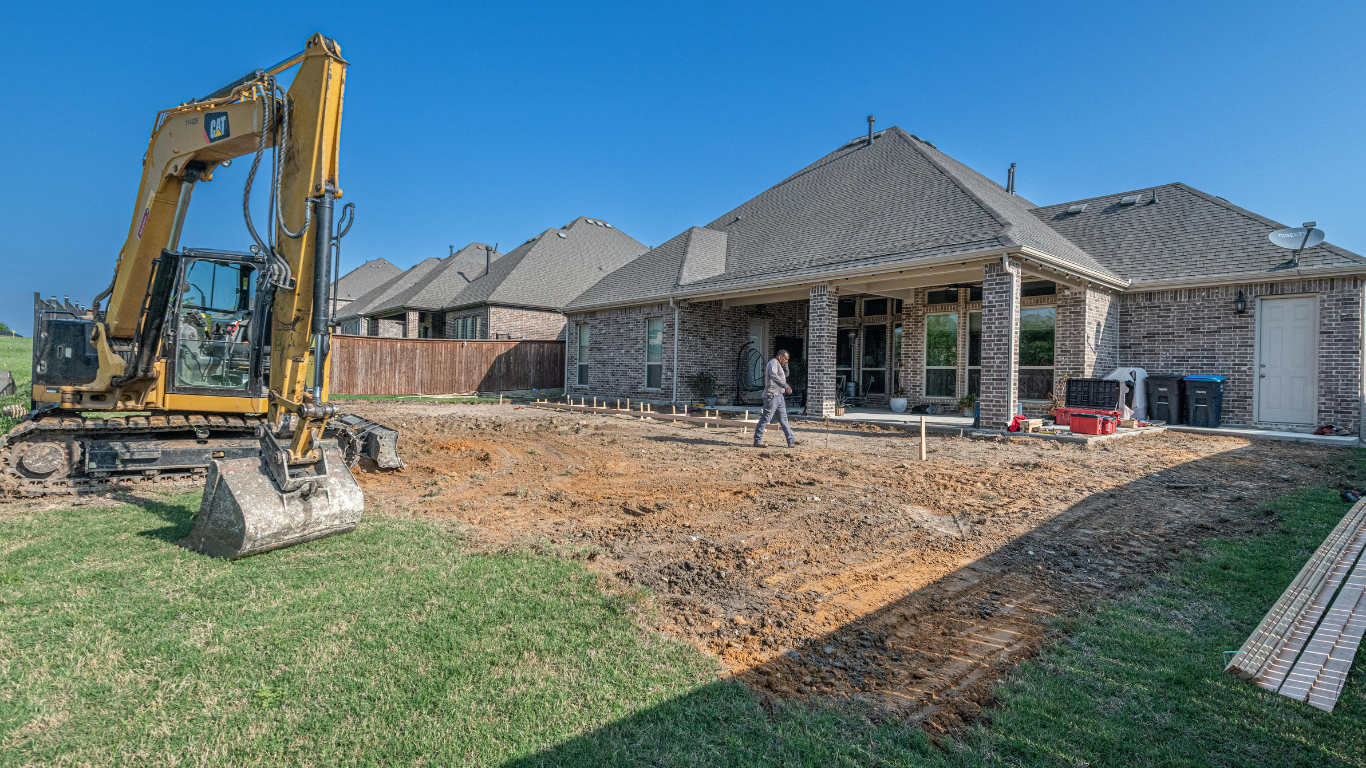 Backyard of a house under construction, showing a large excavator, dirt area being prepared, and a person walking.
