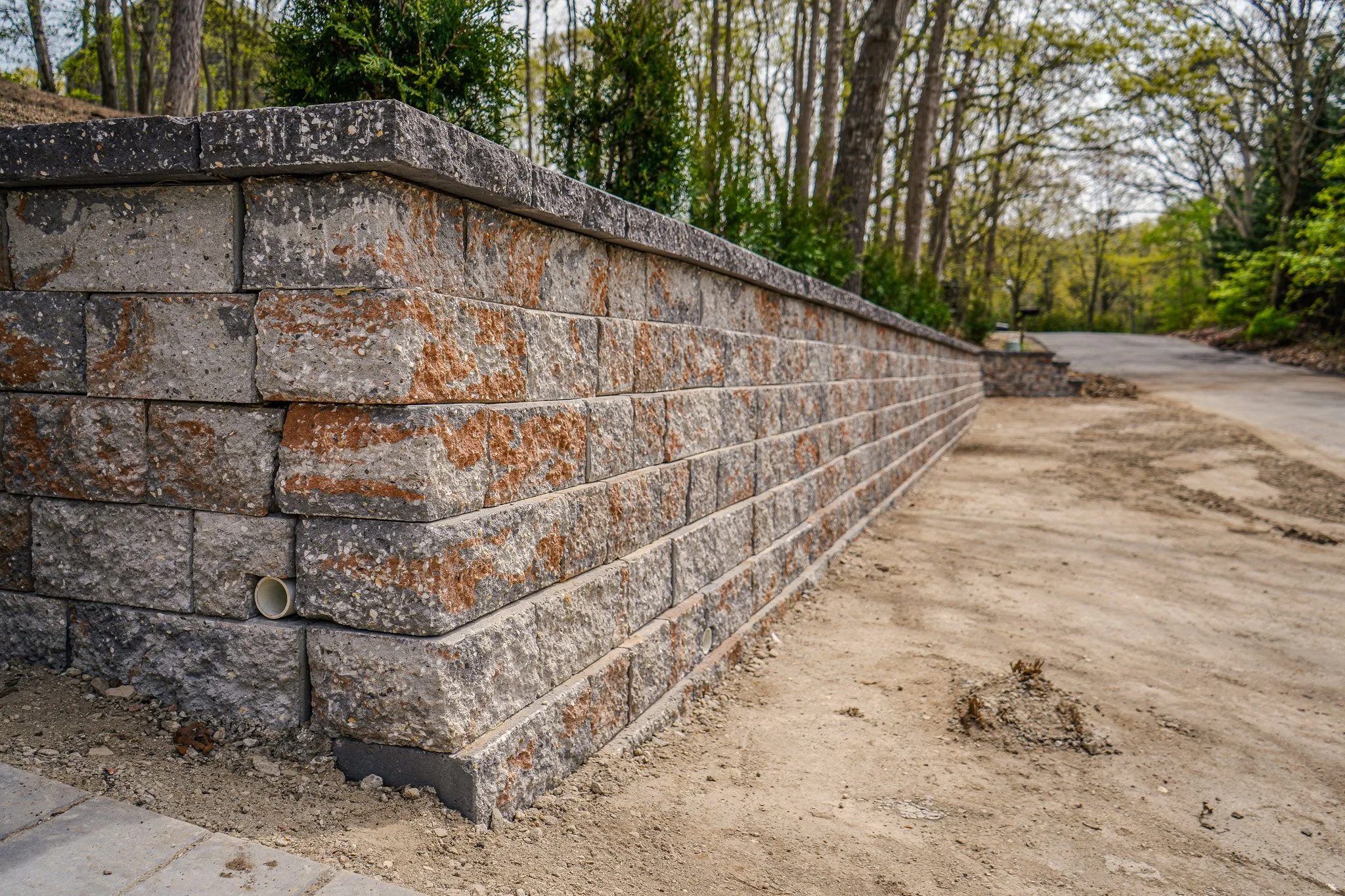 Construction site with a long stone brick retaining wall, dirt pathway, and trees in the background.