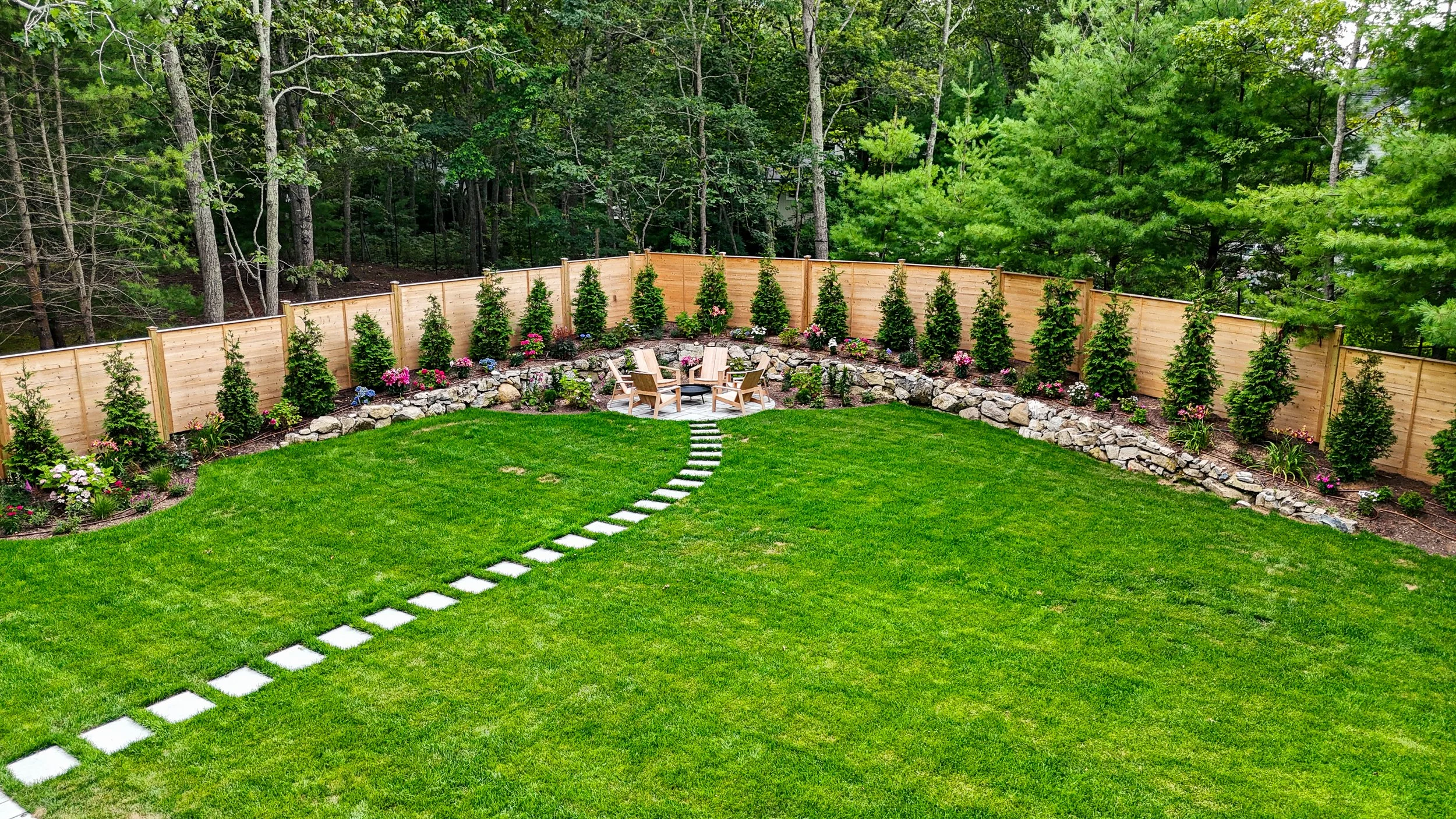 A backyard with a lush green lawn, a curved stone walkway, a wooden fence, tall evergreen trees, and a sitting area with four wooden chairs surrounding a fire pit.
