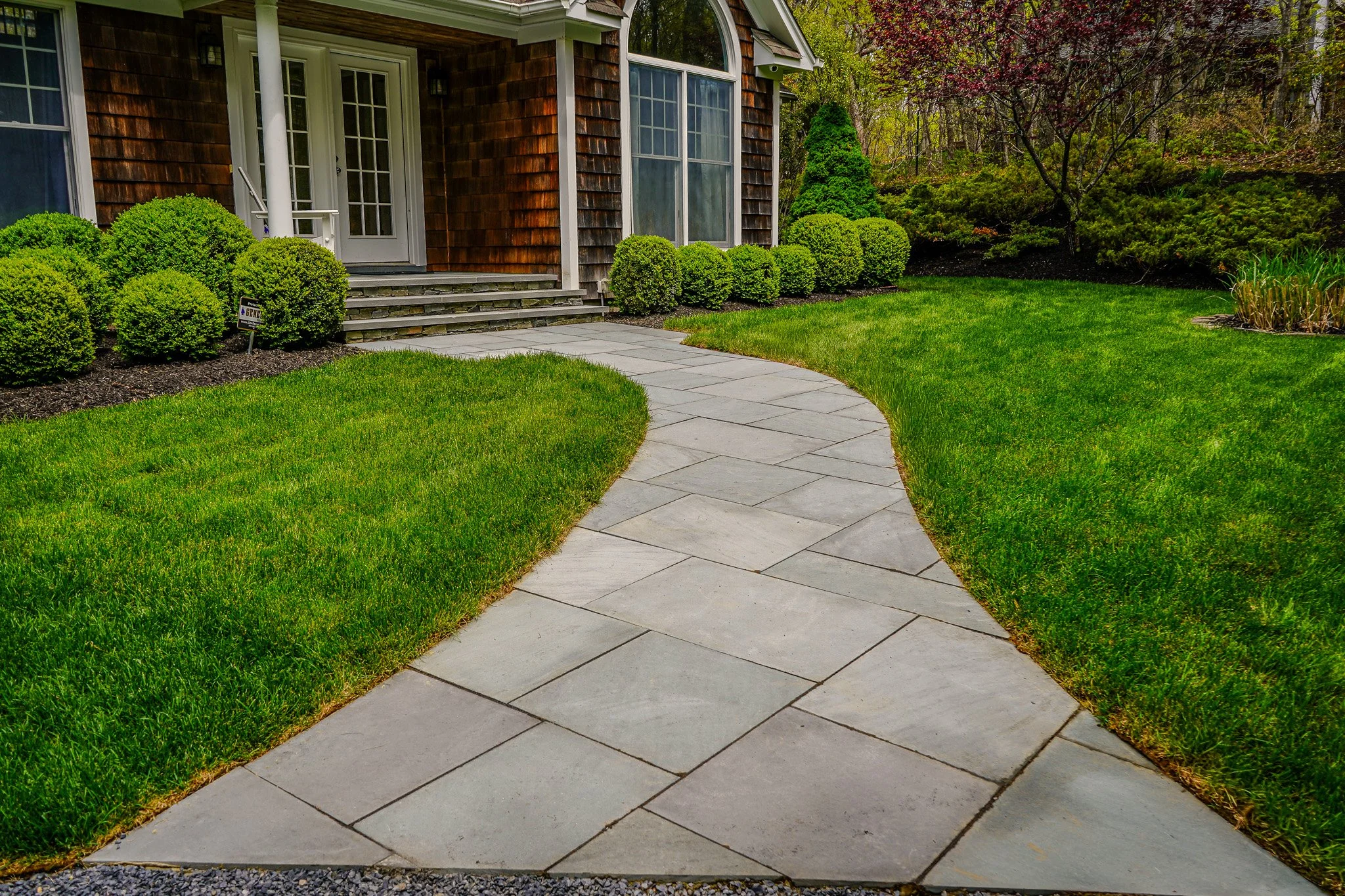 A curved stone pathway leading to the front steps of a house with wooden siding, surrounded by well-manicured green grass and neatly trimmed bushes.