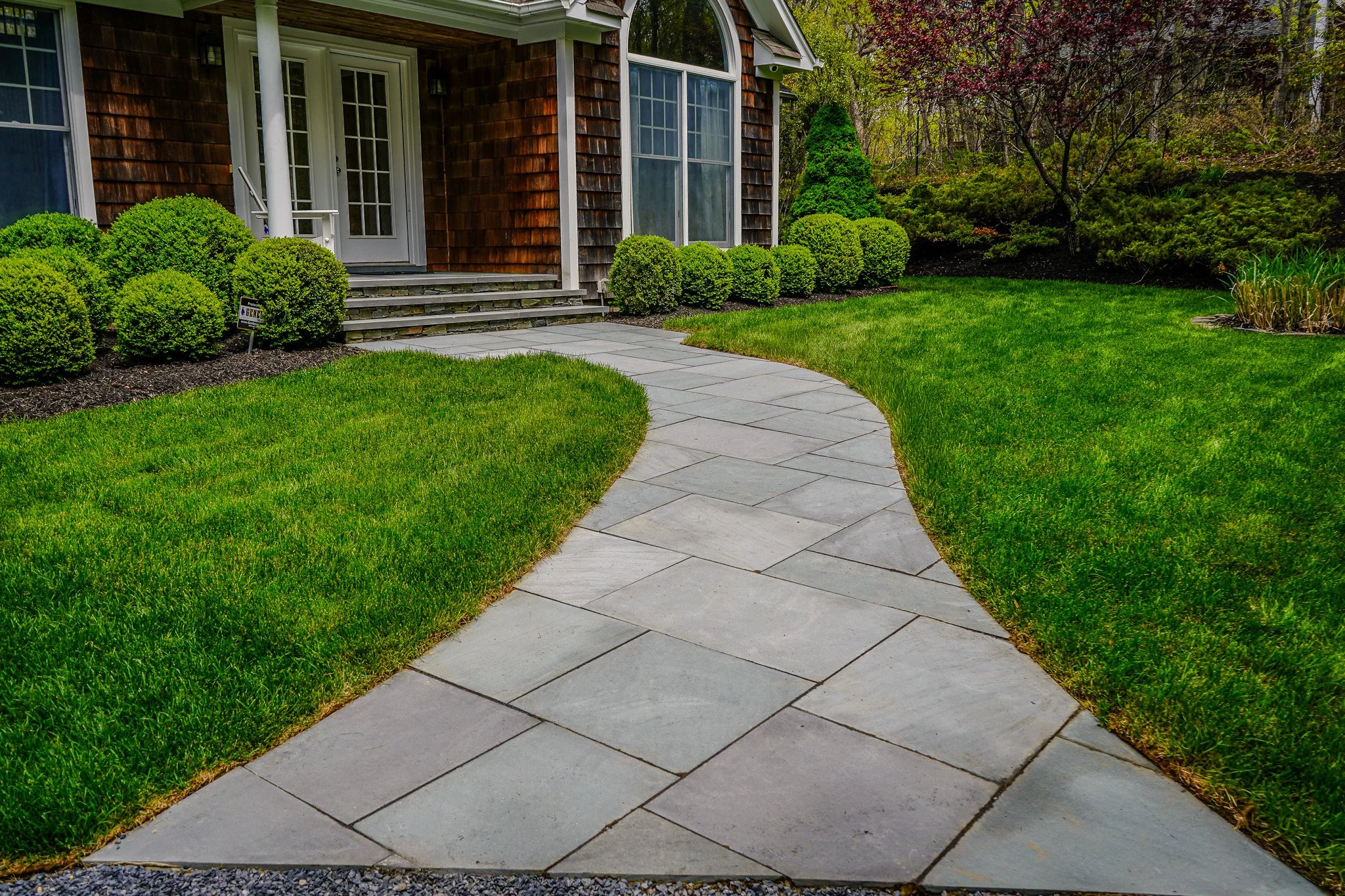 A curved stone pathway leading to a house with a wooden exterior, surrounded by green grass and neatly trimmed bushes.
