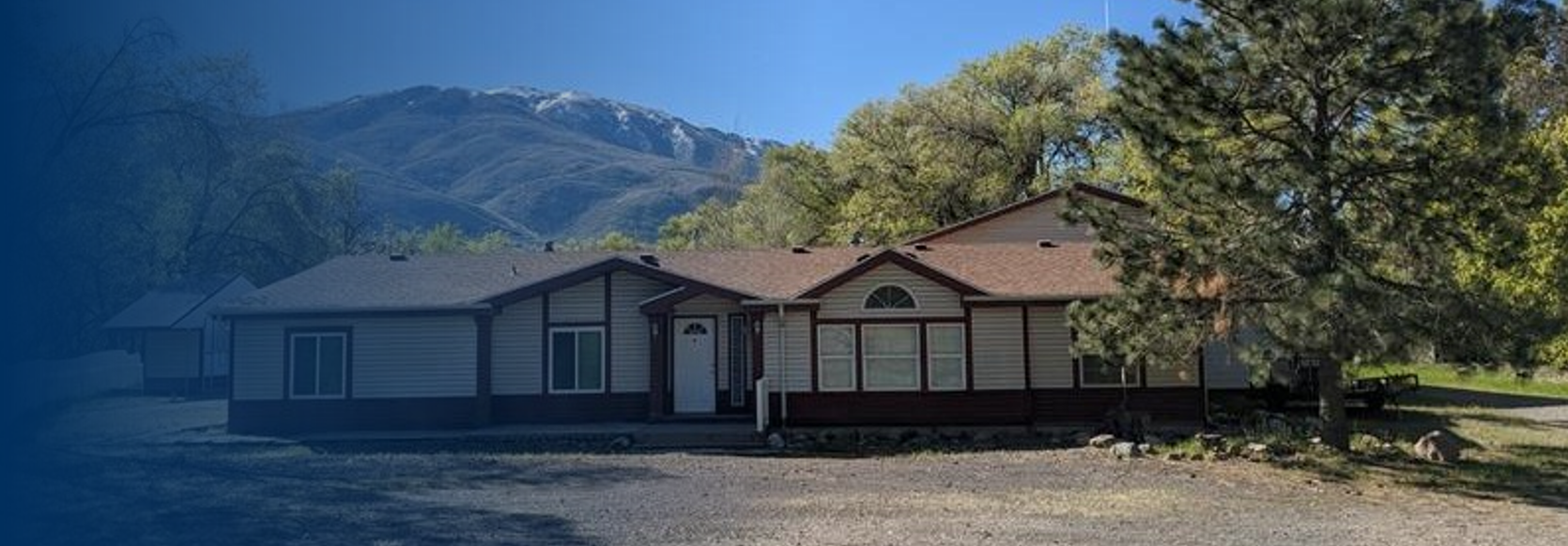 A single-story house with a brown roof and light-colored siding, surrounded by trees and mountains in the background.