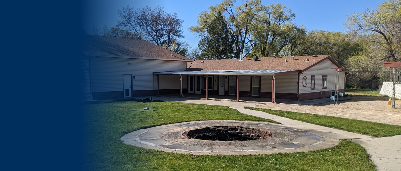 Backyard with a fire pit, green grass, paved patio, and a house with a covered porch and siding, surrounded by trees.