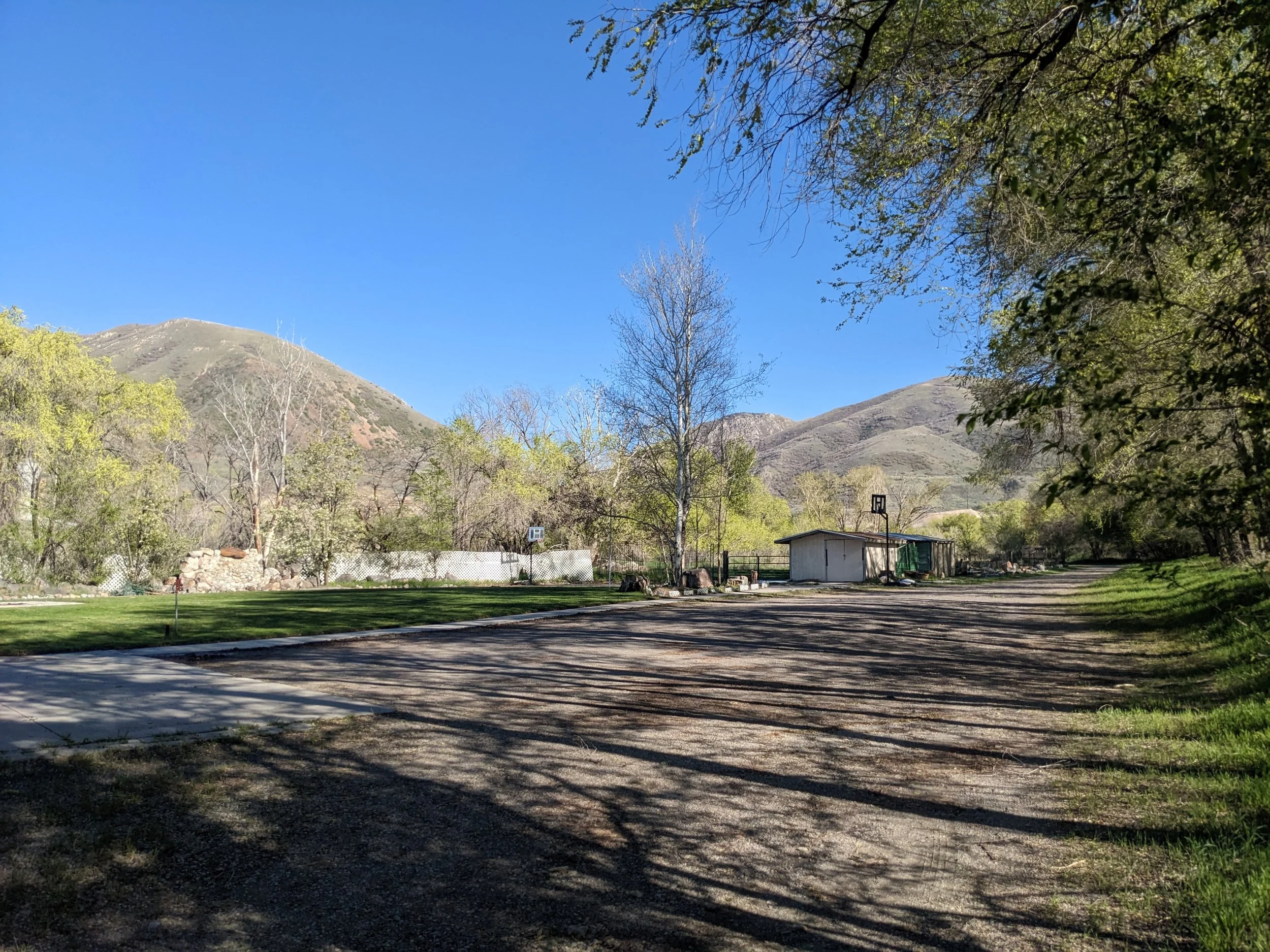 A scenic outdoor scene with a bright blue sky, mountains in the background, and trees with budding leaves. There is a grassy area, a small building, and a basketball hoop on a dirt or gravel area.